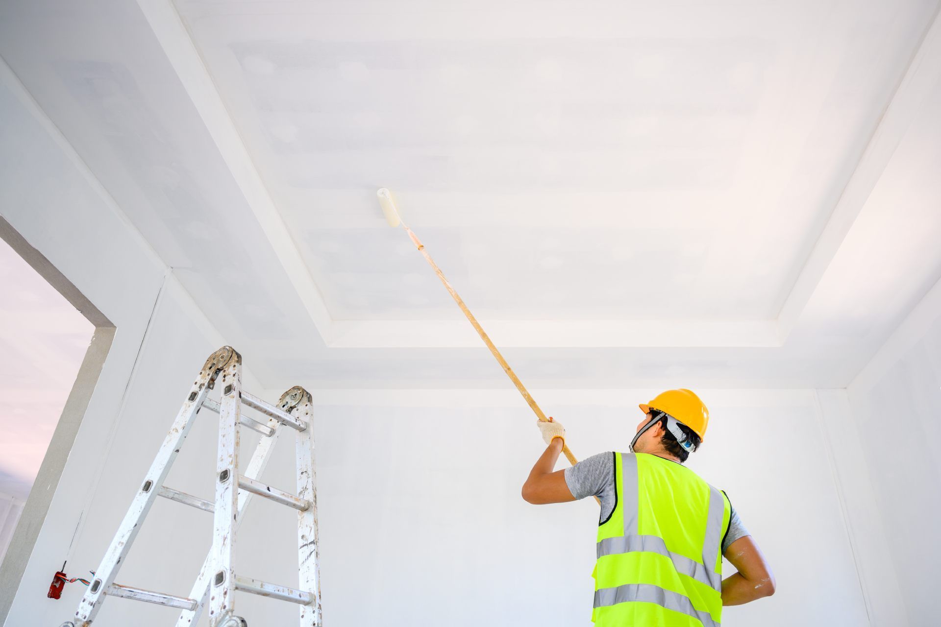 Person in safety vest paints a ceiling white with a roller, using a ladder in a room.