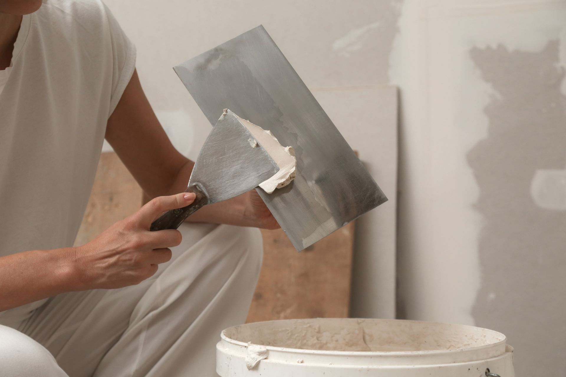 Person in white overalls applying joint compound to a wall with tools.