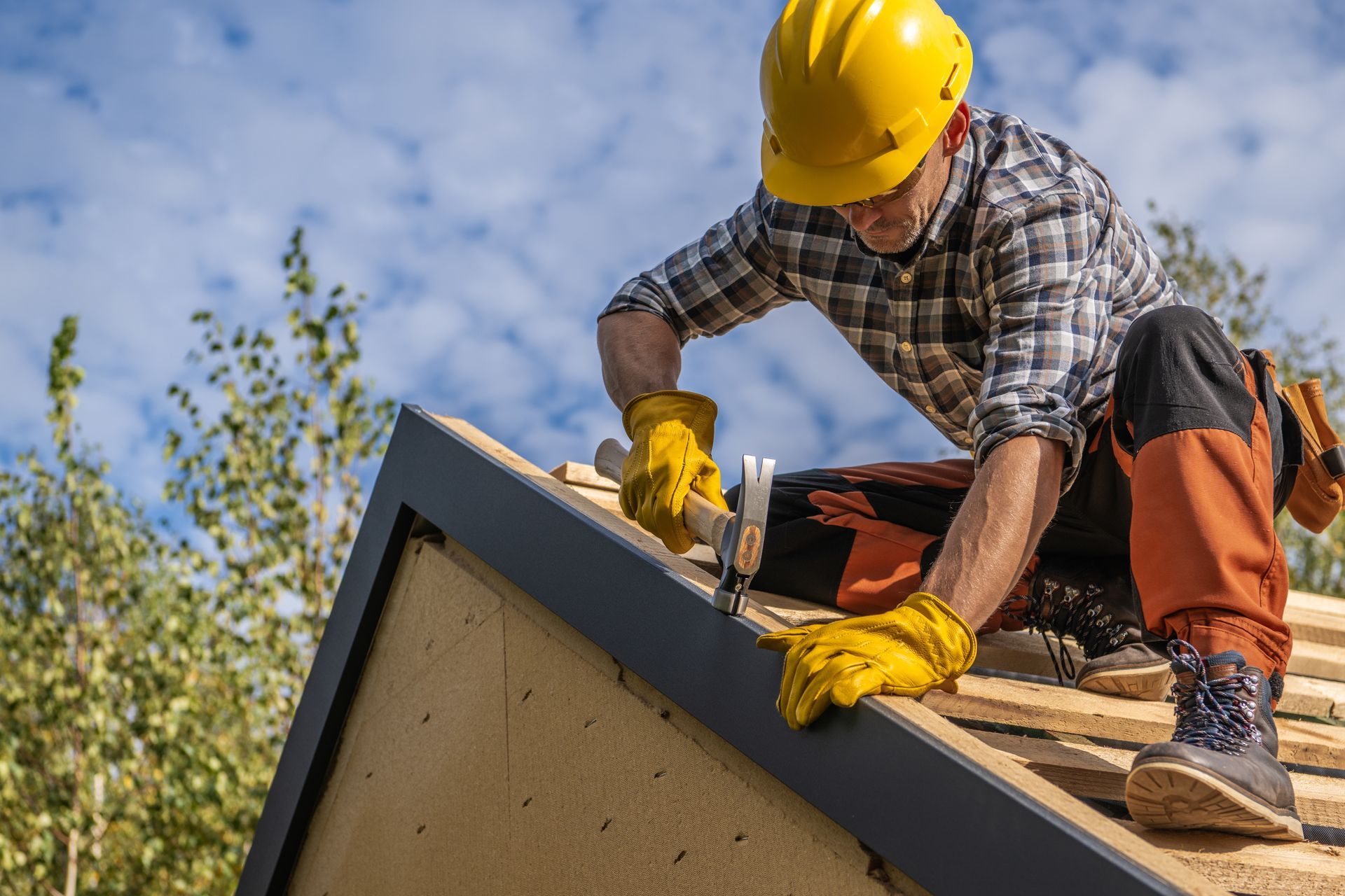 Worker in yellow hard hat and gloves securing shingles on a rooftop under a blue sky
