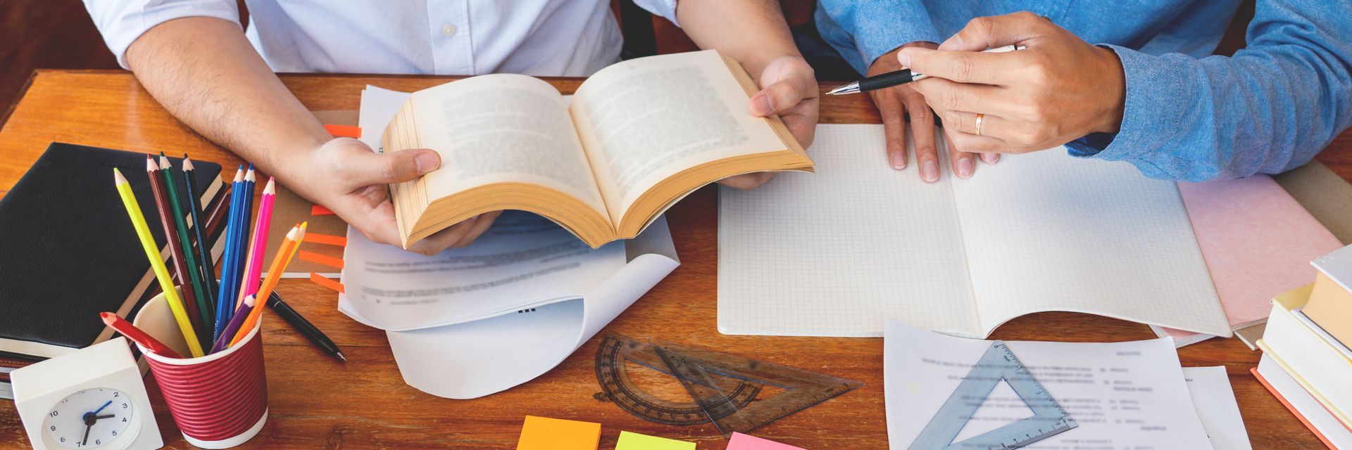 Two people study together, one holding an open book, the other writing in a notebook at a wooden desk.