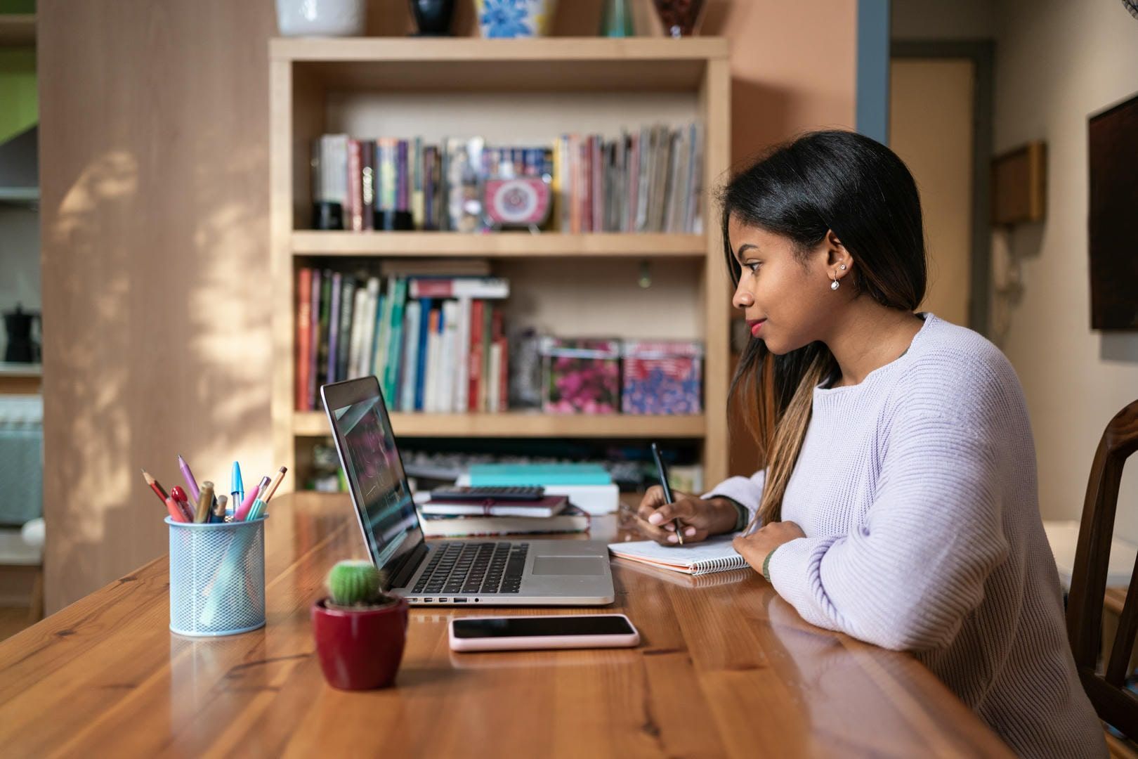 Woman with dark hair writing notes at a wooden table with laptop, books, and pencils.