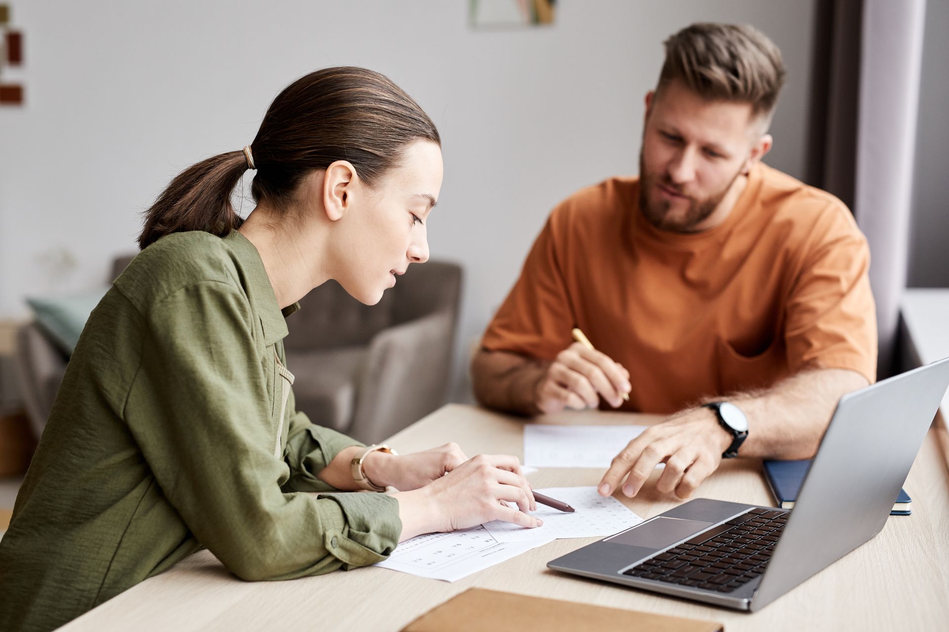 Woman and man reviewing papers at a table with a laptop. Man points with a pen, woman writes, both focused.
