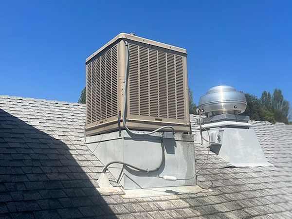 Large, square, weathered air conditioning unit and vent on a gray shingled roof under a clear blue sky.