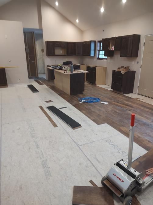 Kitchen under construction: Installing wood flooring with cabinetry in the background. Construction tools and materials visible.