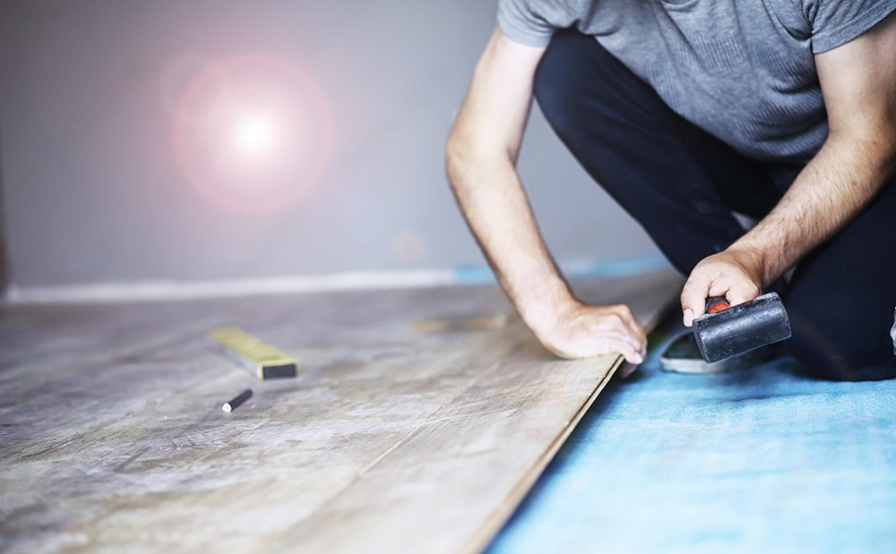 Person installing laminate flooring, kneeling, holding tool, indoor setting.