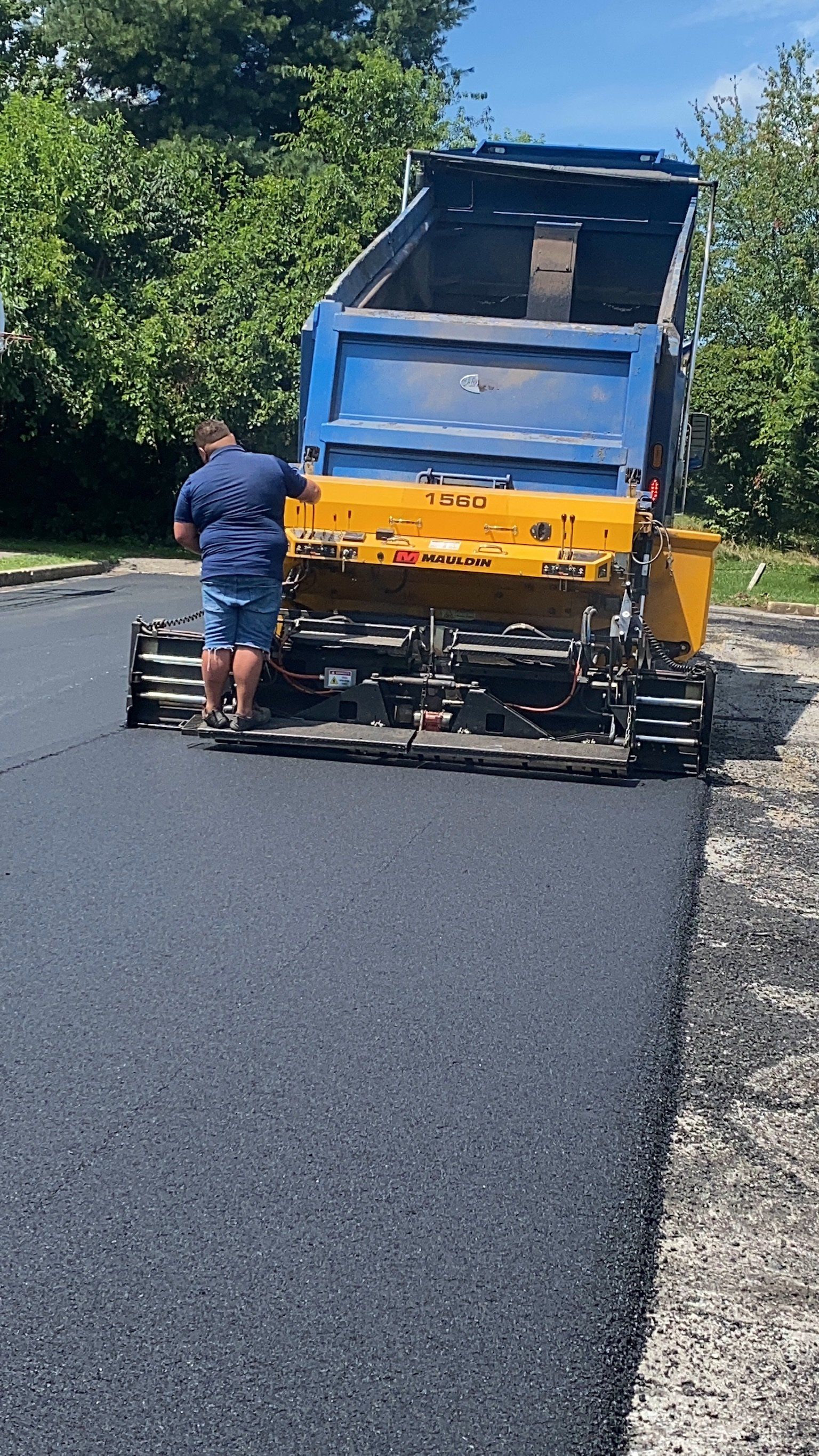 Asphalt paving: A man in shorts operates machinery as a dump truck unloads asphalt onto a road.