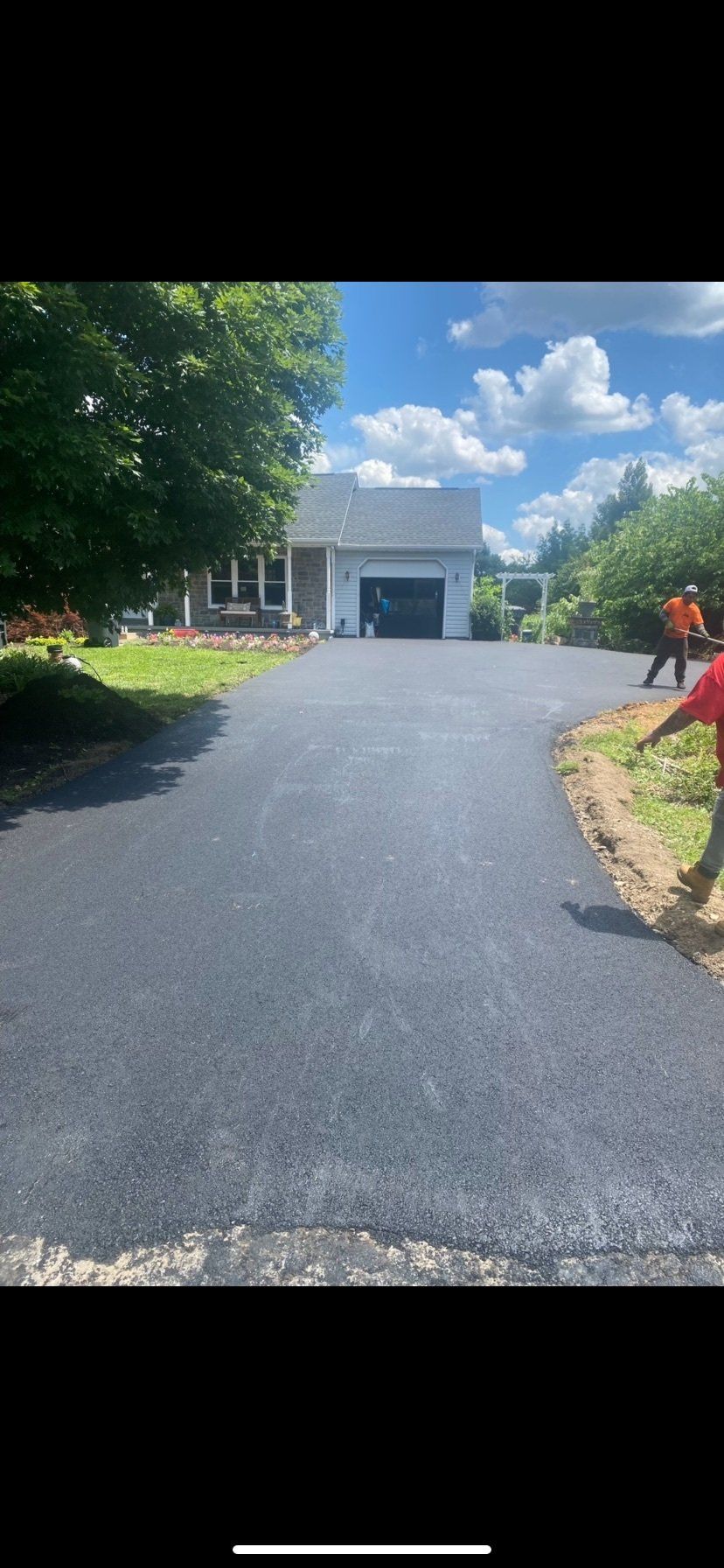 Newly paved driveway leading to a house with a garage, two workers on the side, blue sky with clouds.