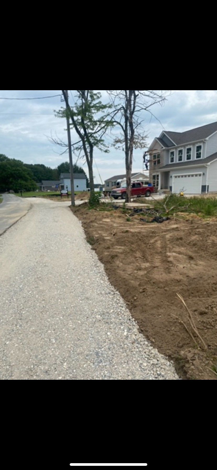 A gravel road next to dirt with houses and trees in the background under a cloudy sky.