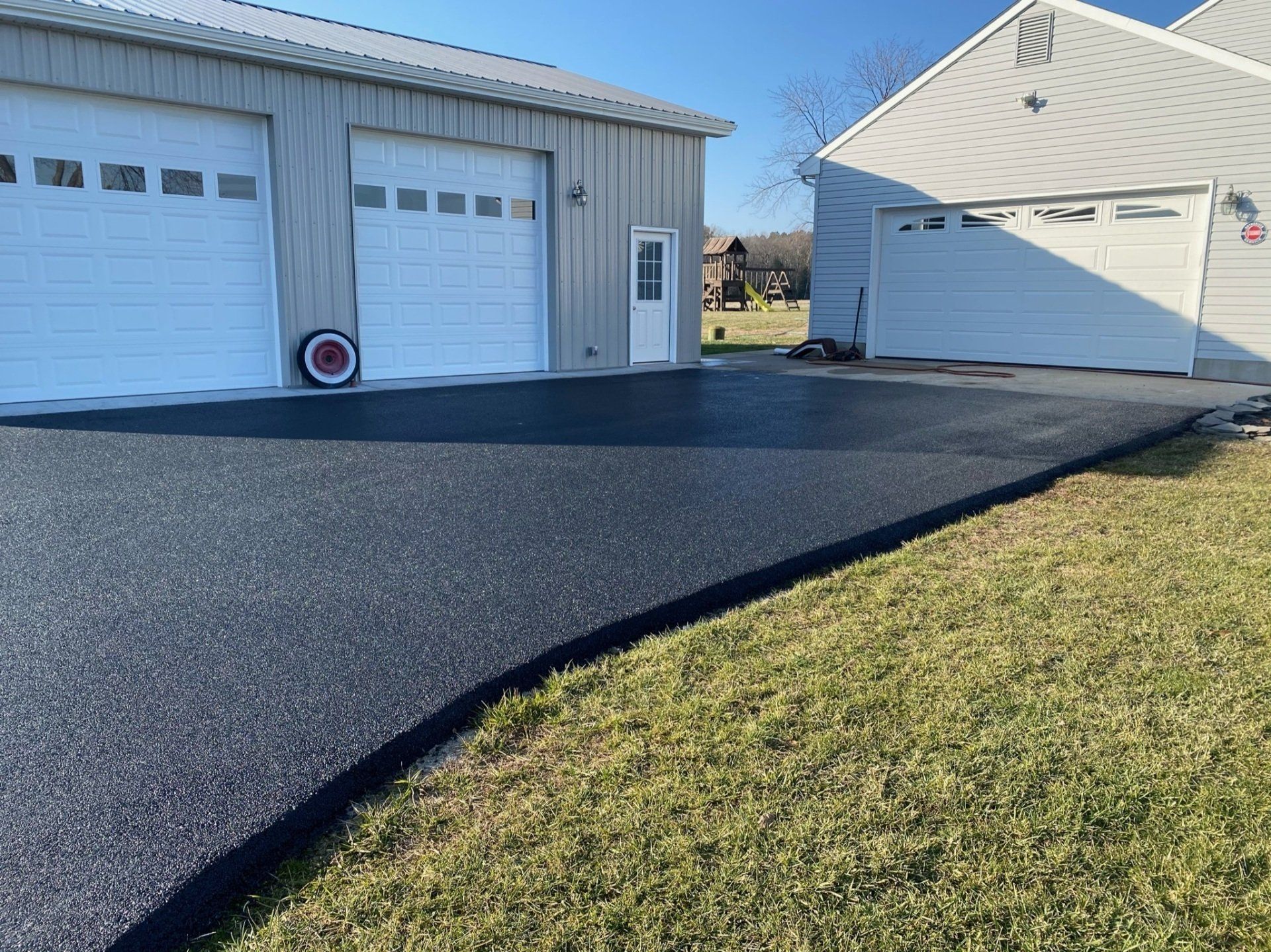 Newly paved black asphalt driveway next to a grassy yard and two white garage buildings.