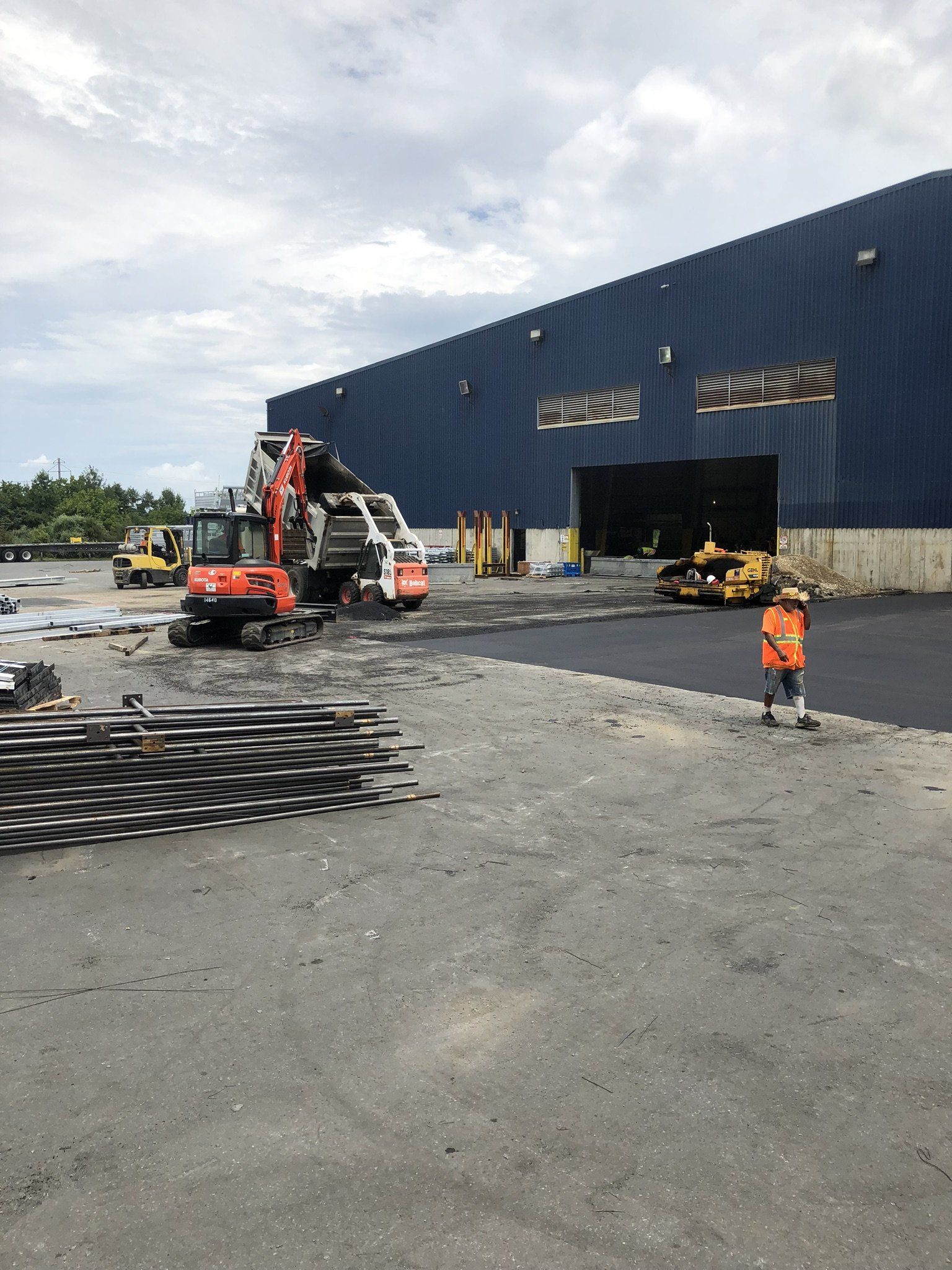Construction site. Excavator, worker, and equipment near blue building. Piles of metal in foreground, asphalt ground.