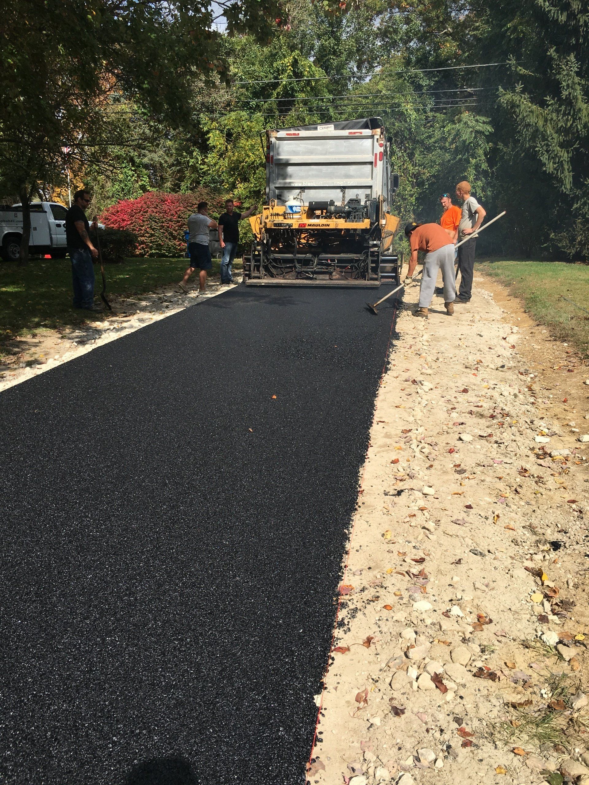 Asphalt paving in progress: machine laying blacktop on a dirt road, workers raking it smooth.