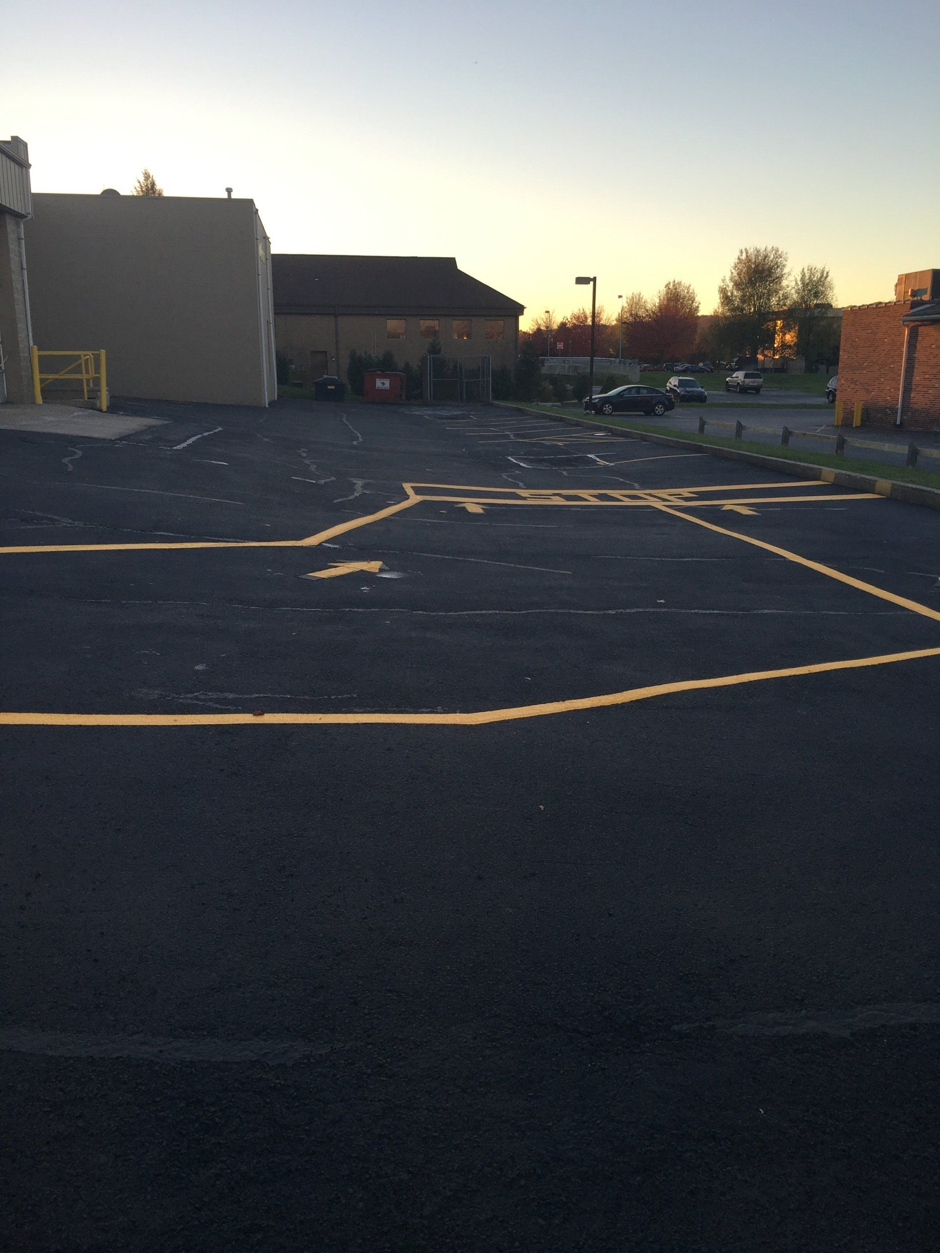 Paved parking lot with yellow lines, buildings, trees, and cars in the distance. Dusk setting with a clear sky.