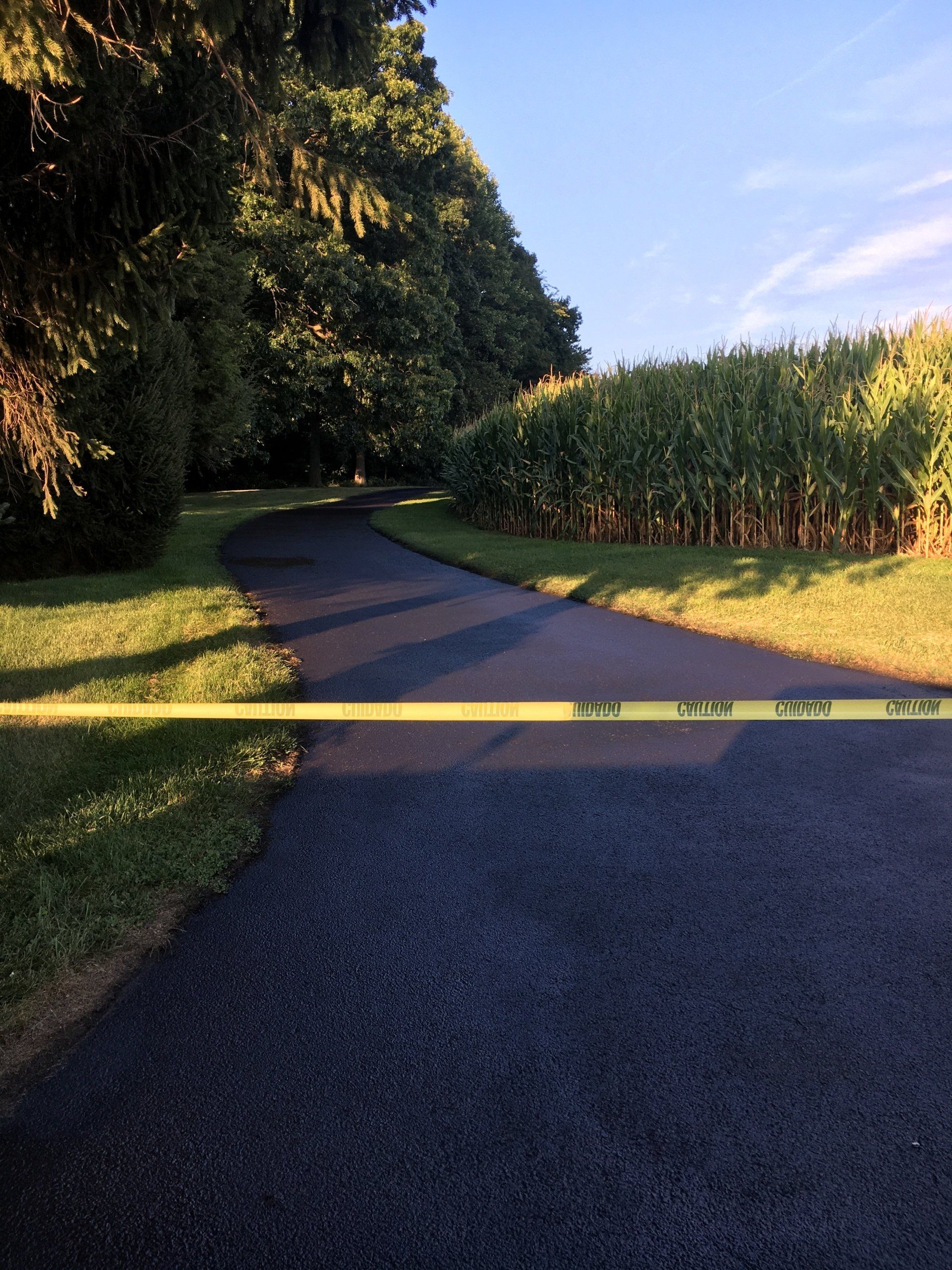 Asphalt road blocked by yellow caution tape, leading uphill past green trees and a field of crops.