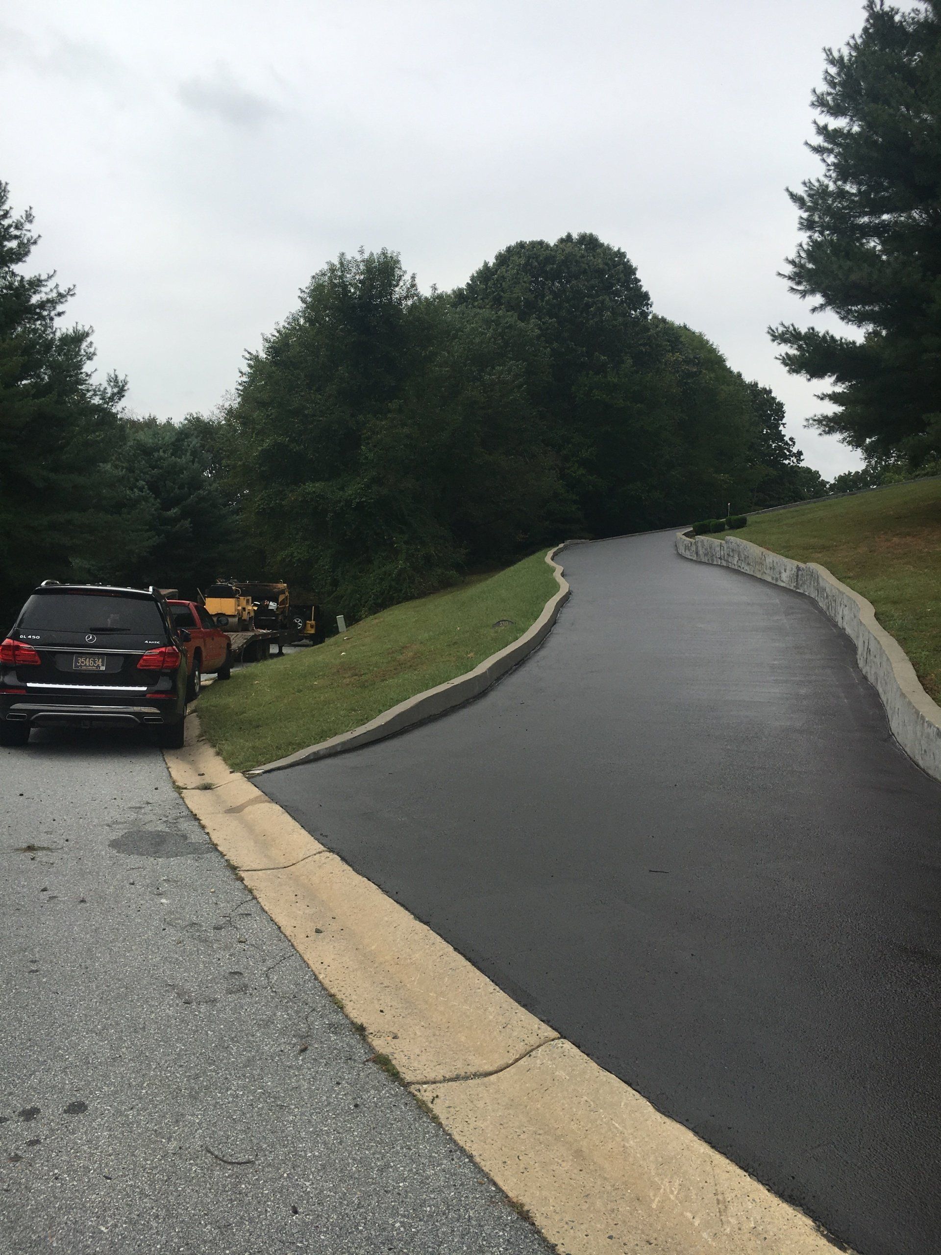 Black asphalt driveway curves uphill, bordered by green grass and a concrete curb. A vehicle is parked on gravel at the left.