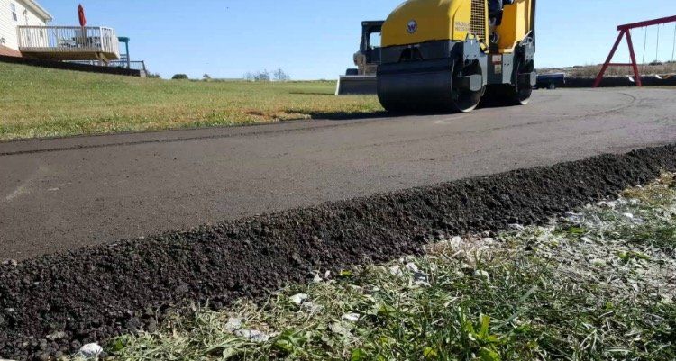 Asphalt paving: A yellow roller compacts fresh black asphalt on a new path, grassy background, blue sky.