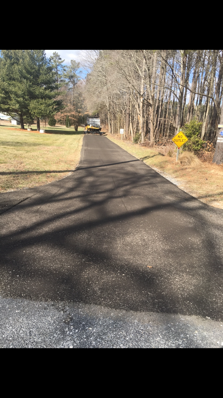 Newly paved asphalt driveway leading toward trees; yellow caution sign on right.