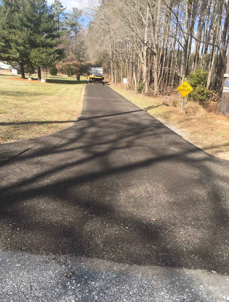 Newly paved dark asphalt driveway leads to a truck. Trees line both sides of the road.