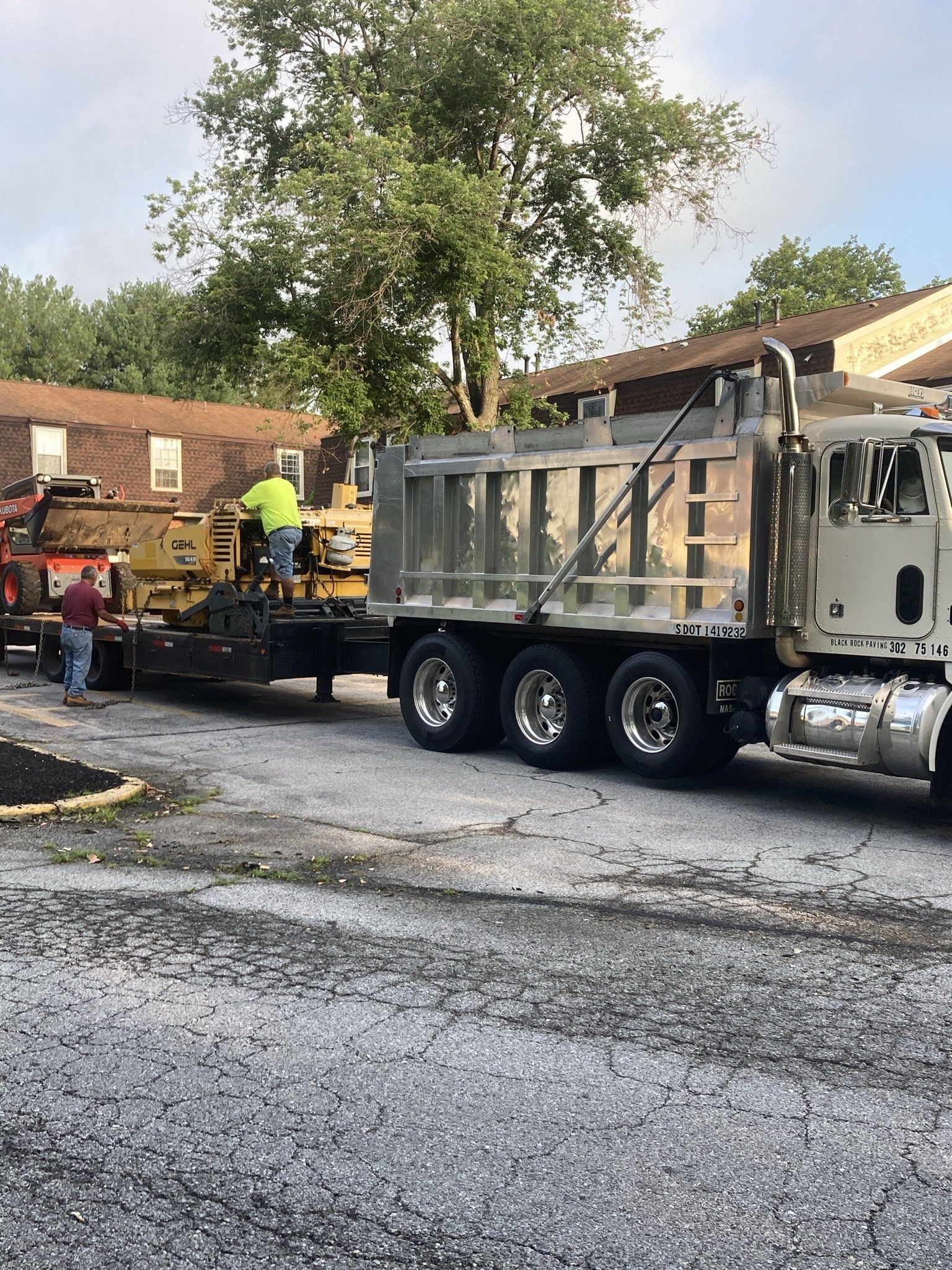 Truck with a flatbed trailer carrying construction equipment, with workers in a parking lot.