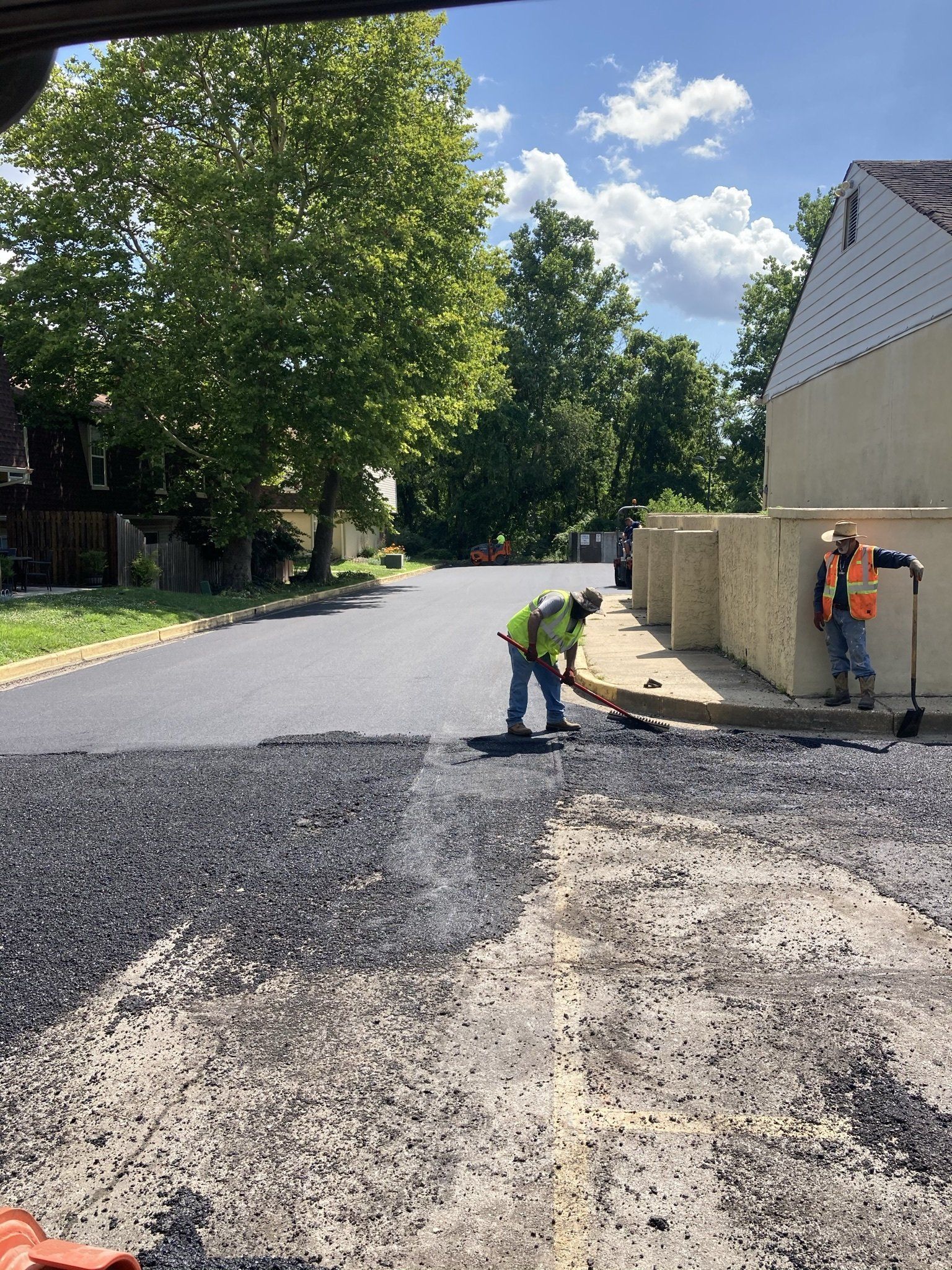 Workers paving a street; one rakes asphalt, the other watches. Sunny day, residential area.