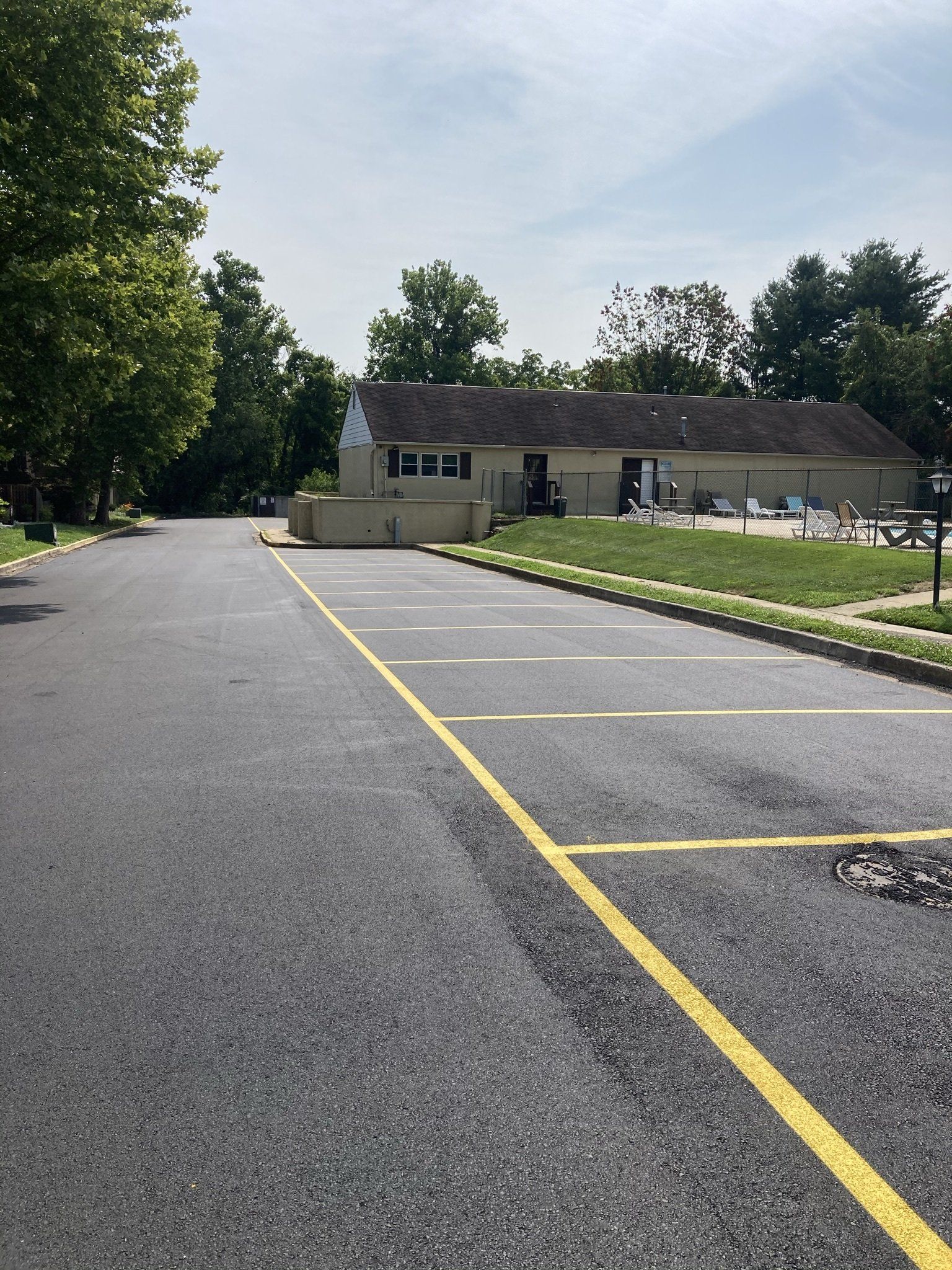 Paved parking lot with yellow lines, building in background, trees on the left, blue sky.