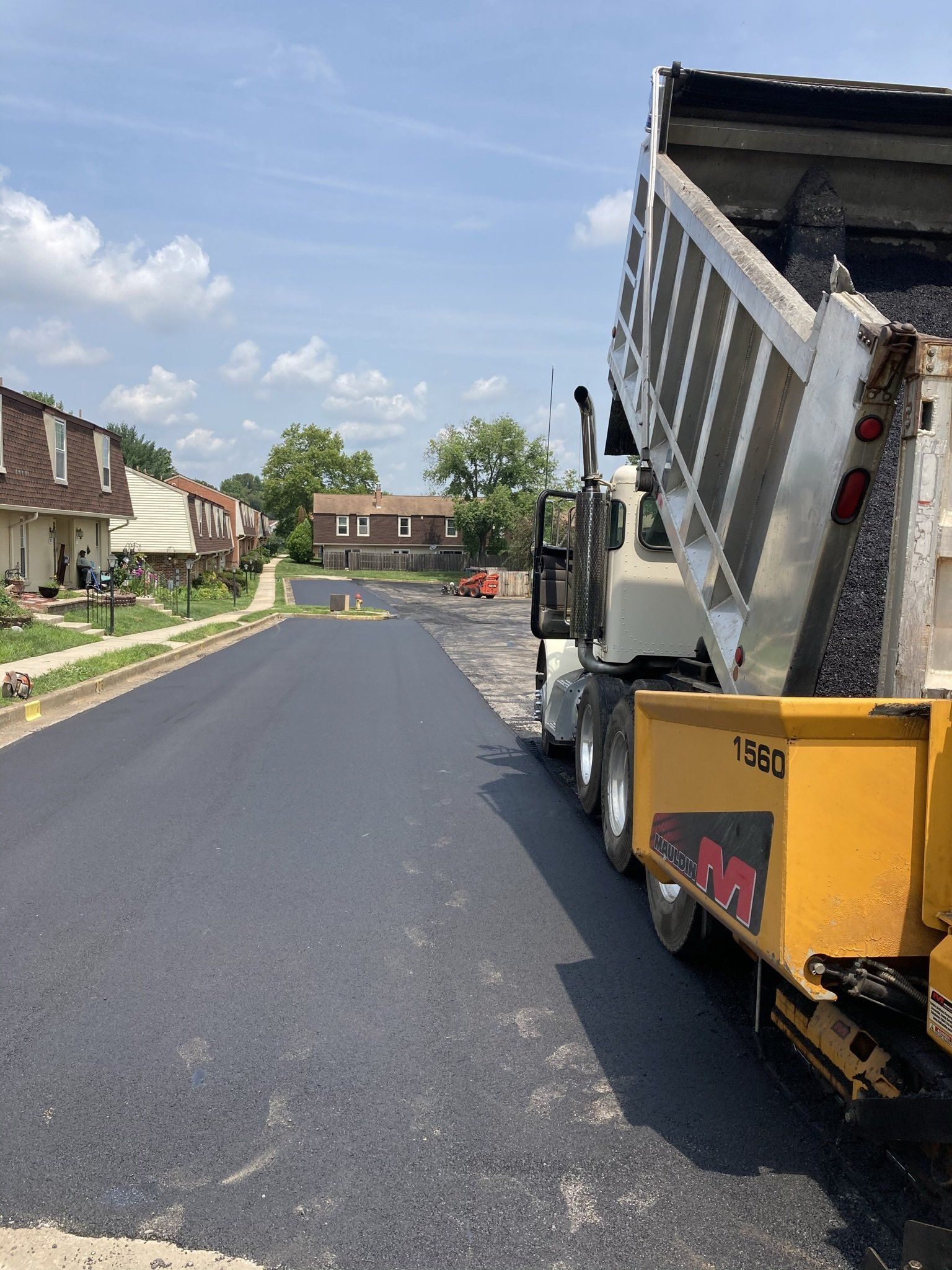 Asphalt paving with a dump truck unloading material on a sunny day. Houses line the street.