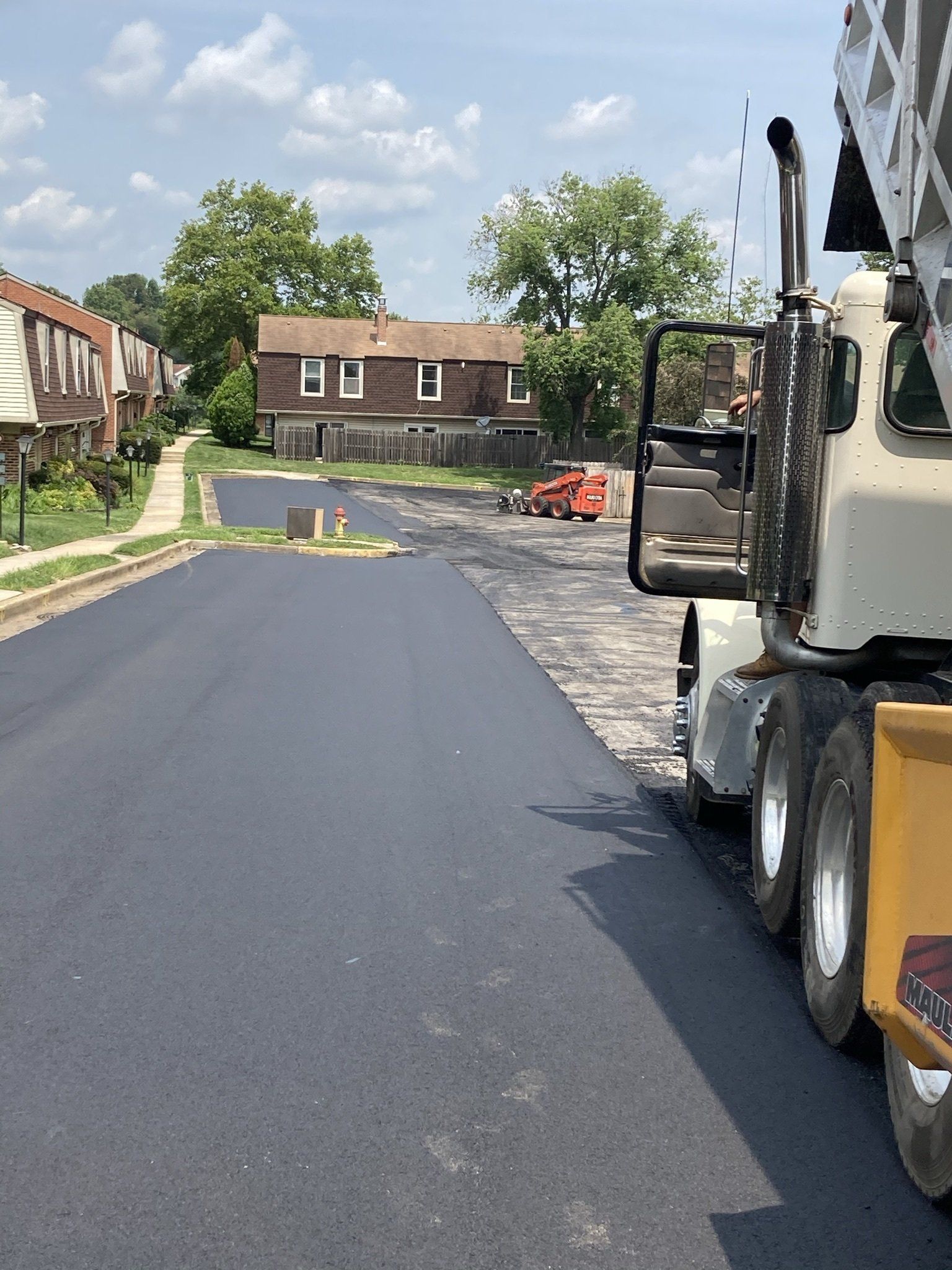 Asphalt paving street with construction truck, residential homes in background.