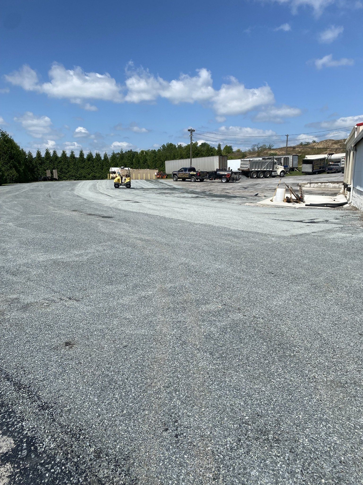 Gravel lot with vehicles, trees in the distance, and a blue sky with white clouds.