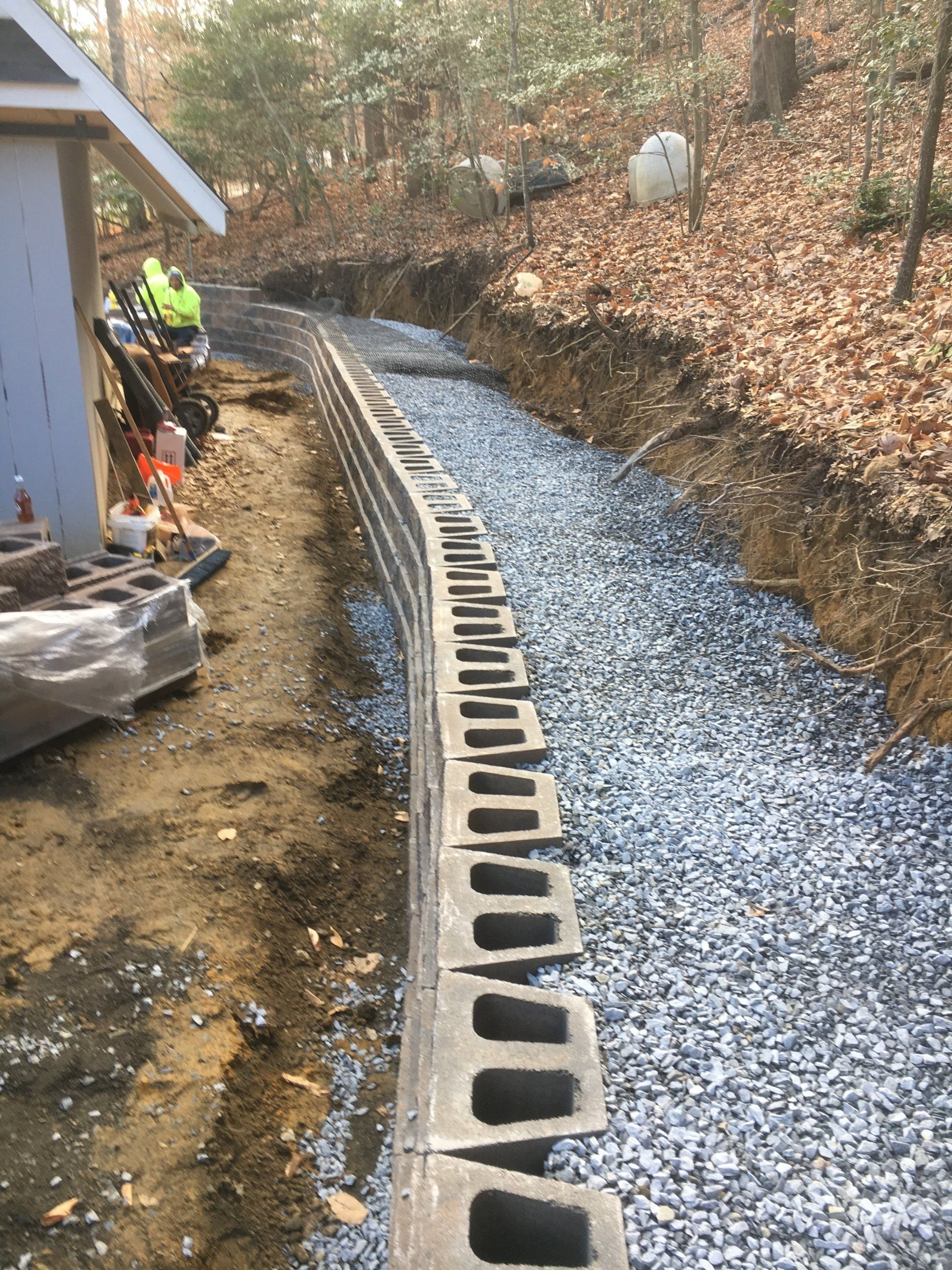 Retaining wall construction with gray blocks and gravel, near a building and woods.