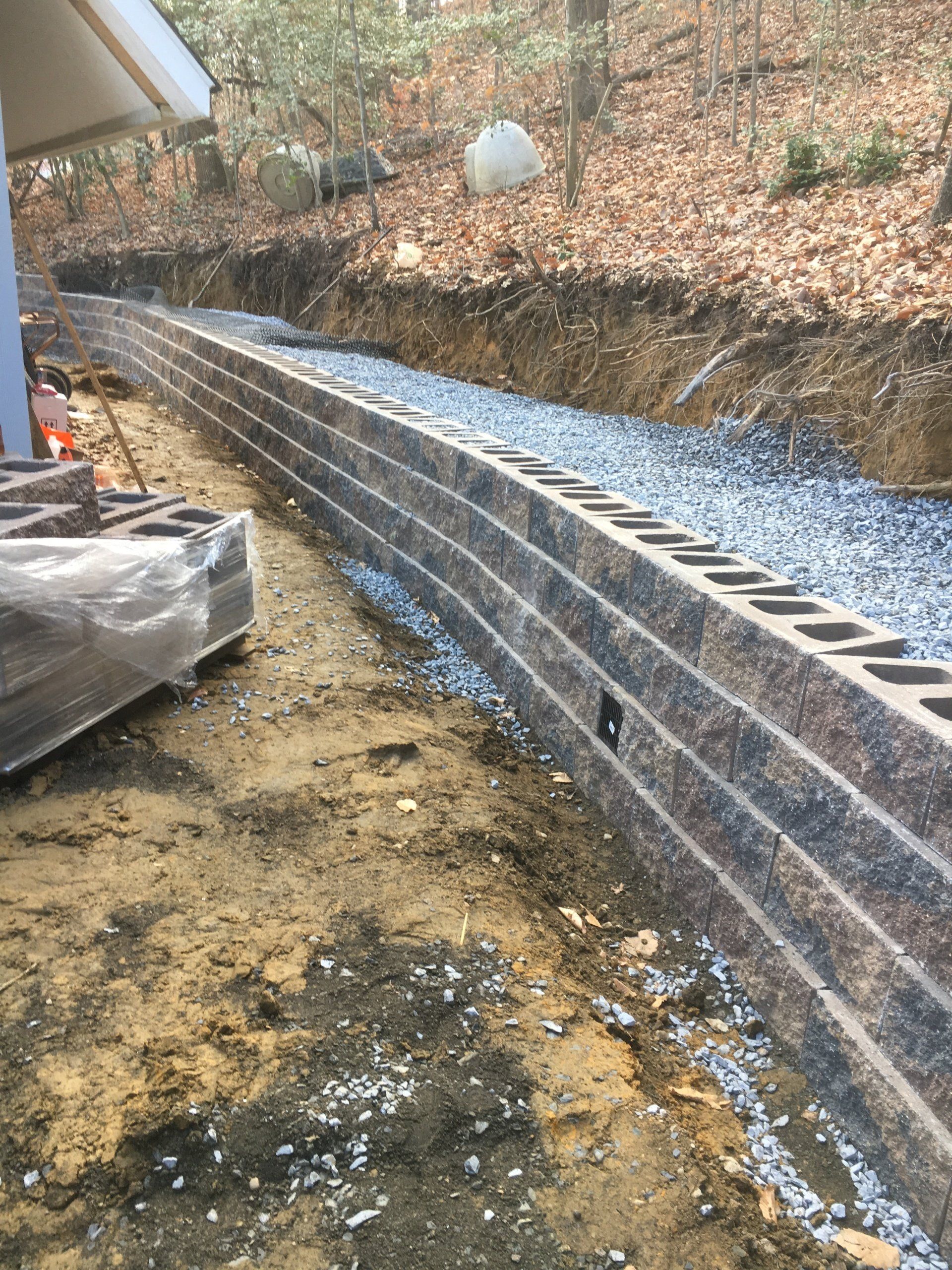 Retaining wall under construction with dark blocks, gravel, and earthen bank.