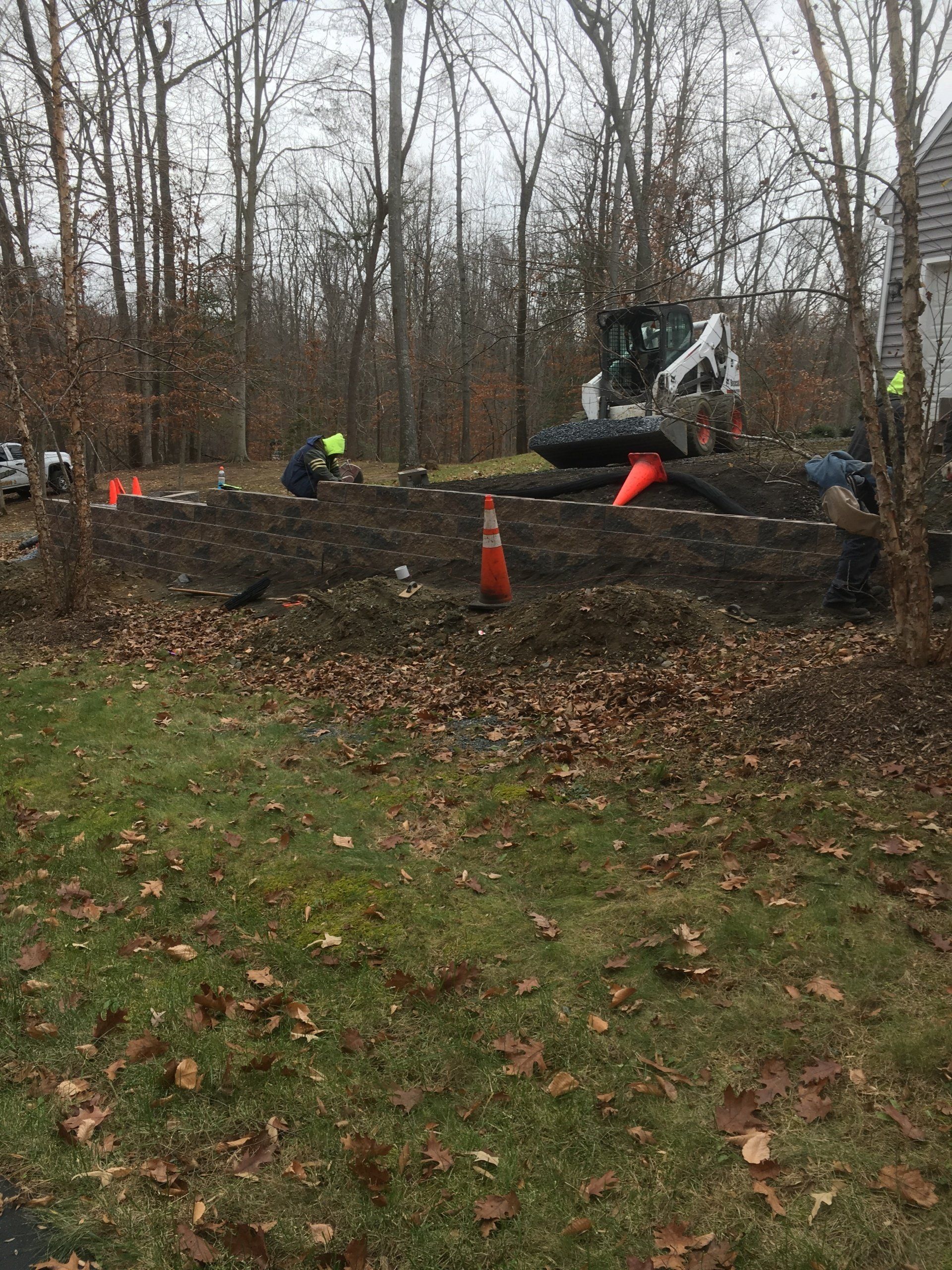 Construction site with a Bobcat excavator, orange cones, and workers near trees.