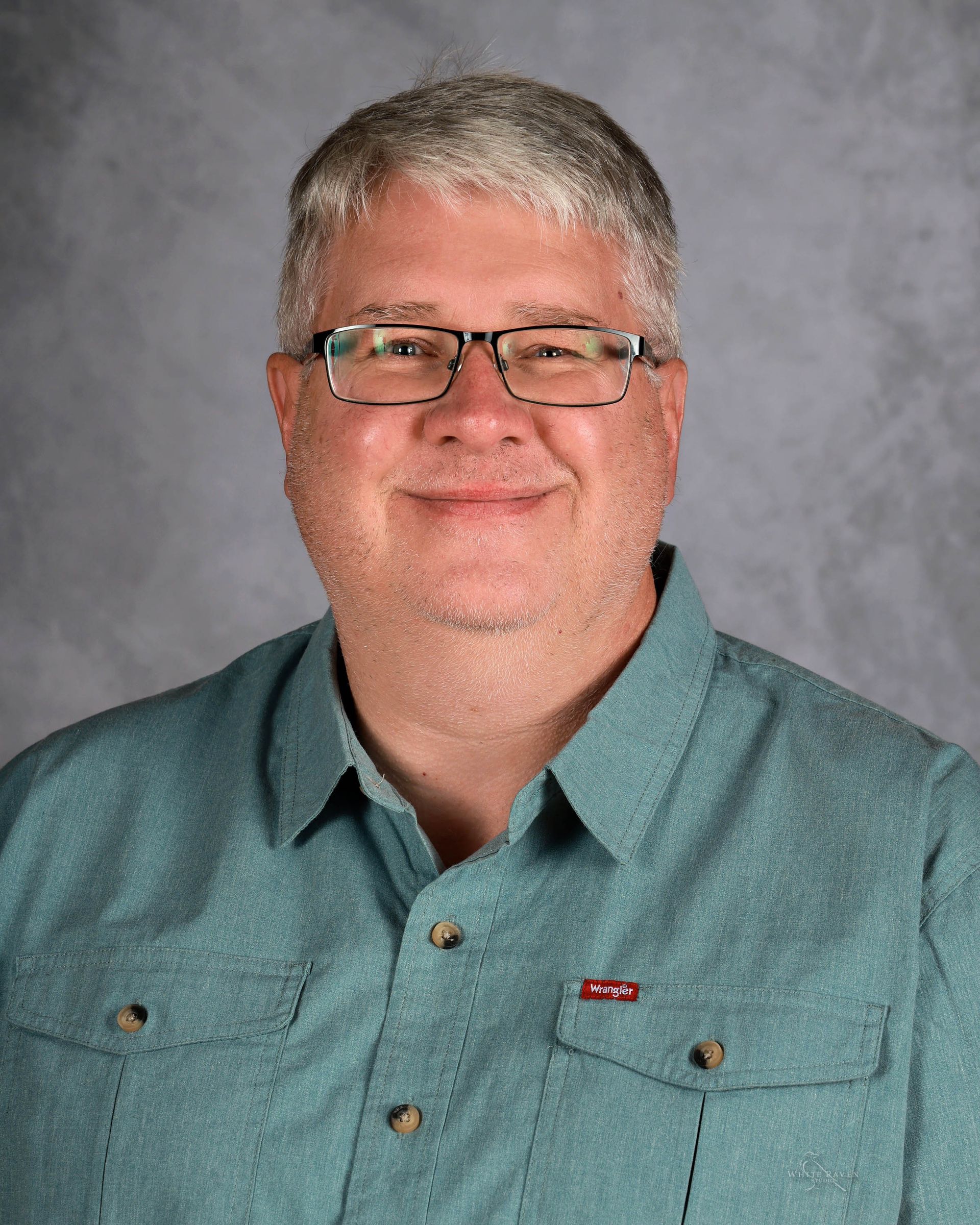 Man with glasses, gray hair, in a teal shirt, smiling, against a gray backdrop.