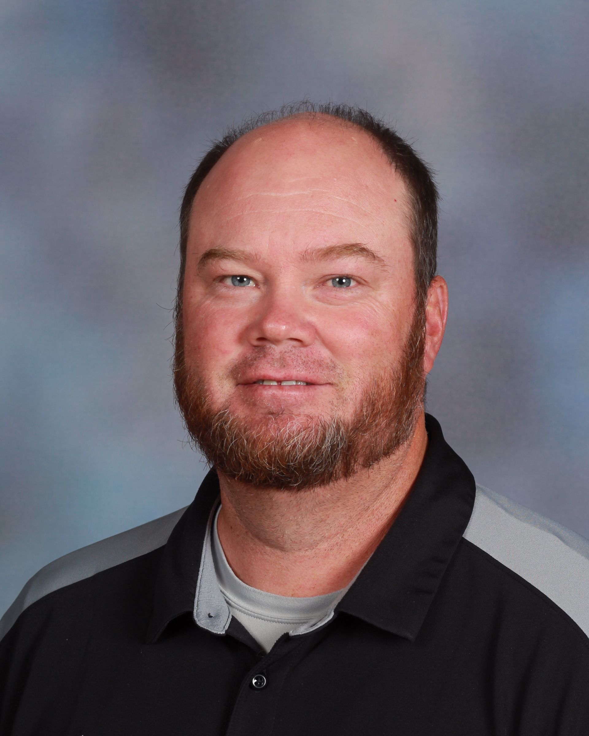 Man with a beard smiling, wearing a black and gray shirt, set against a blurred background.