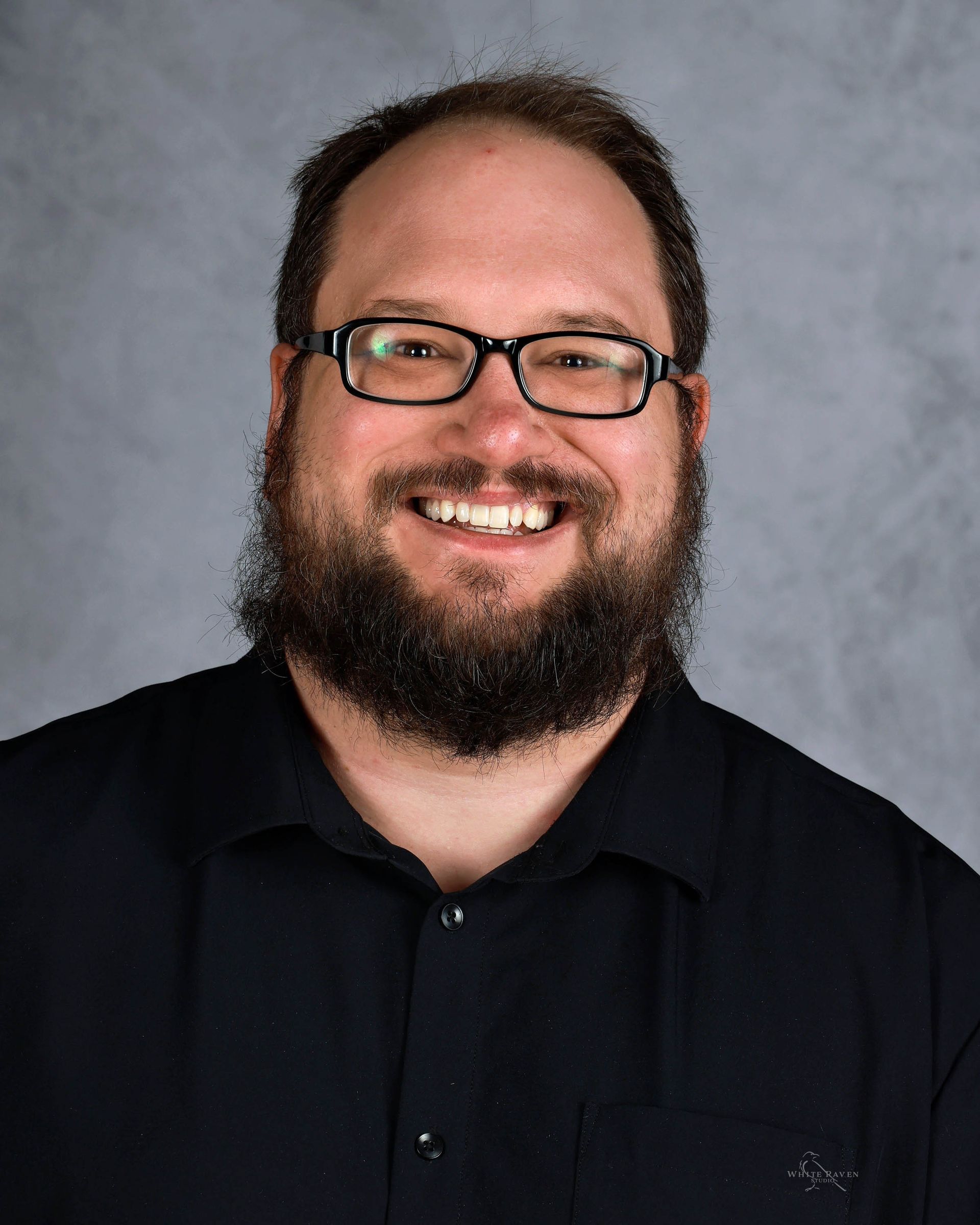 Smiling man with glasses and full beard, wearing a black shirt, against a gray background.