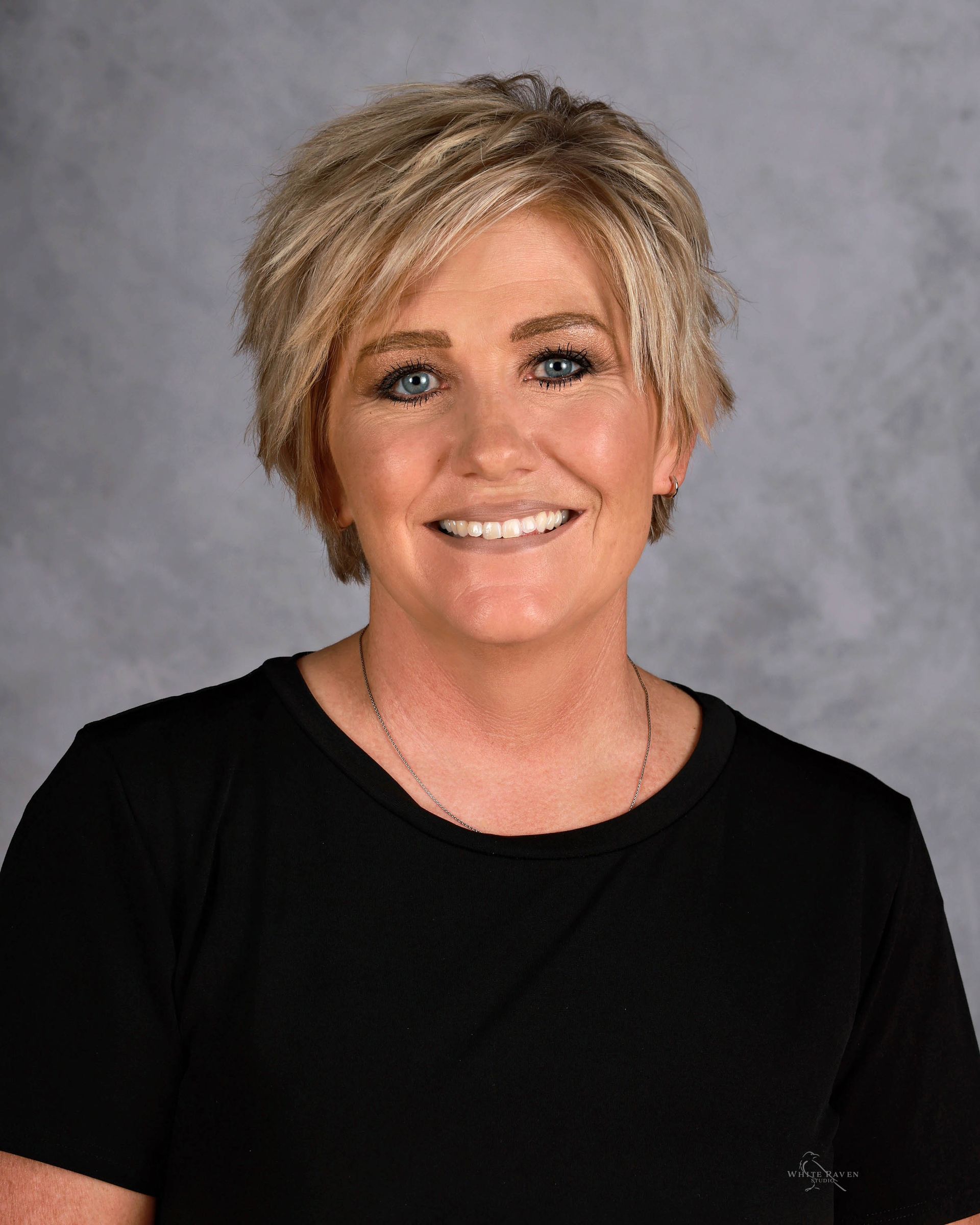 Woman with blonde short hair smiles, wearing a black shirt against a grey background.
