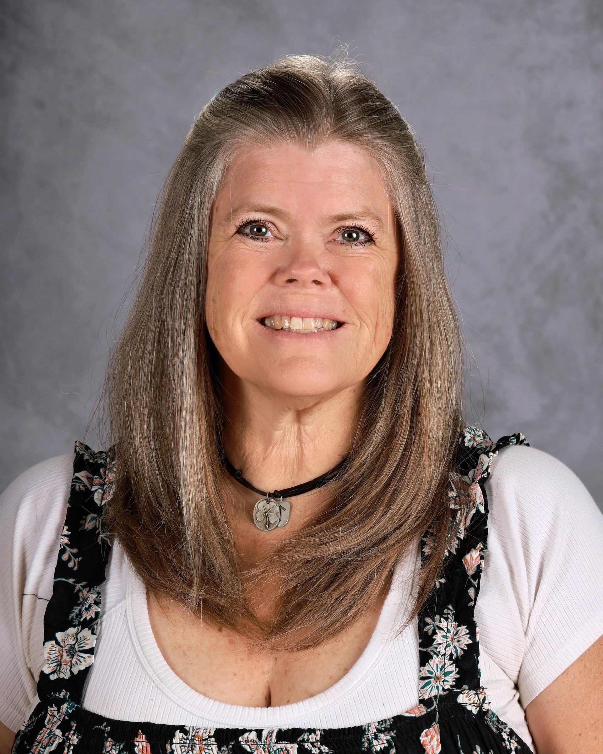 Woman with long gray hair, smiling, wearing a floral patterned top over a white shirt.