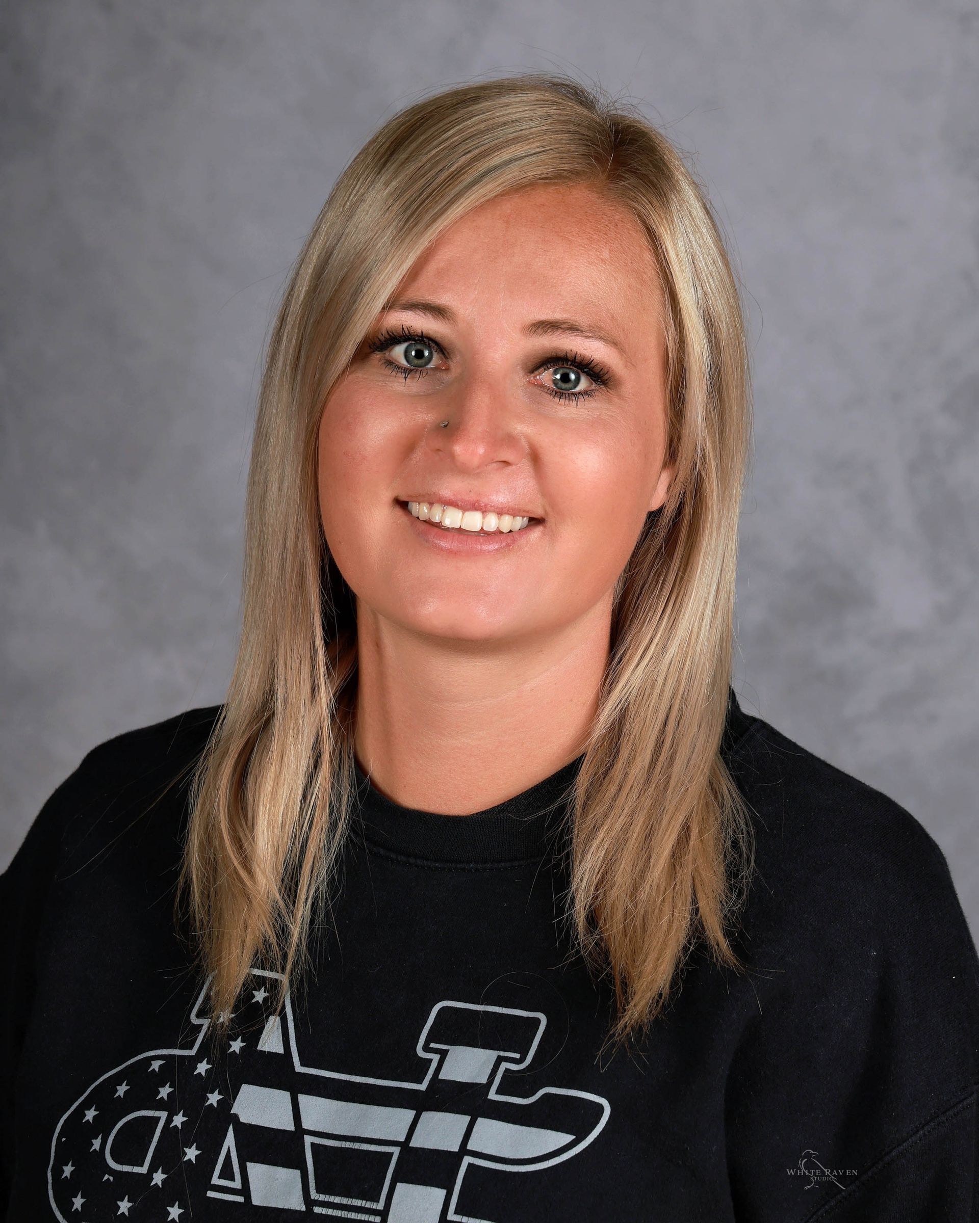 Blonde woman smiling at the camera, wearing a black sweatshirt with a design on it. Gray background.