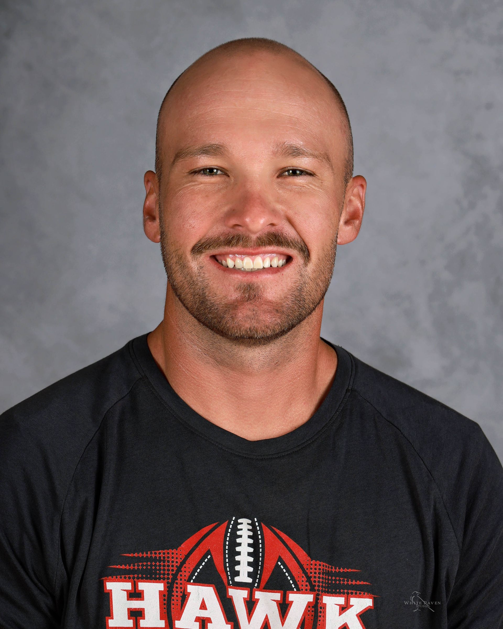 Bald man with a slight smile, wearing a black t-shirt with a red and white football graphic that says