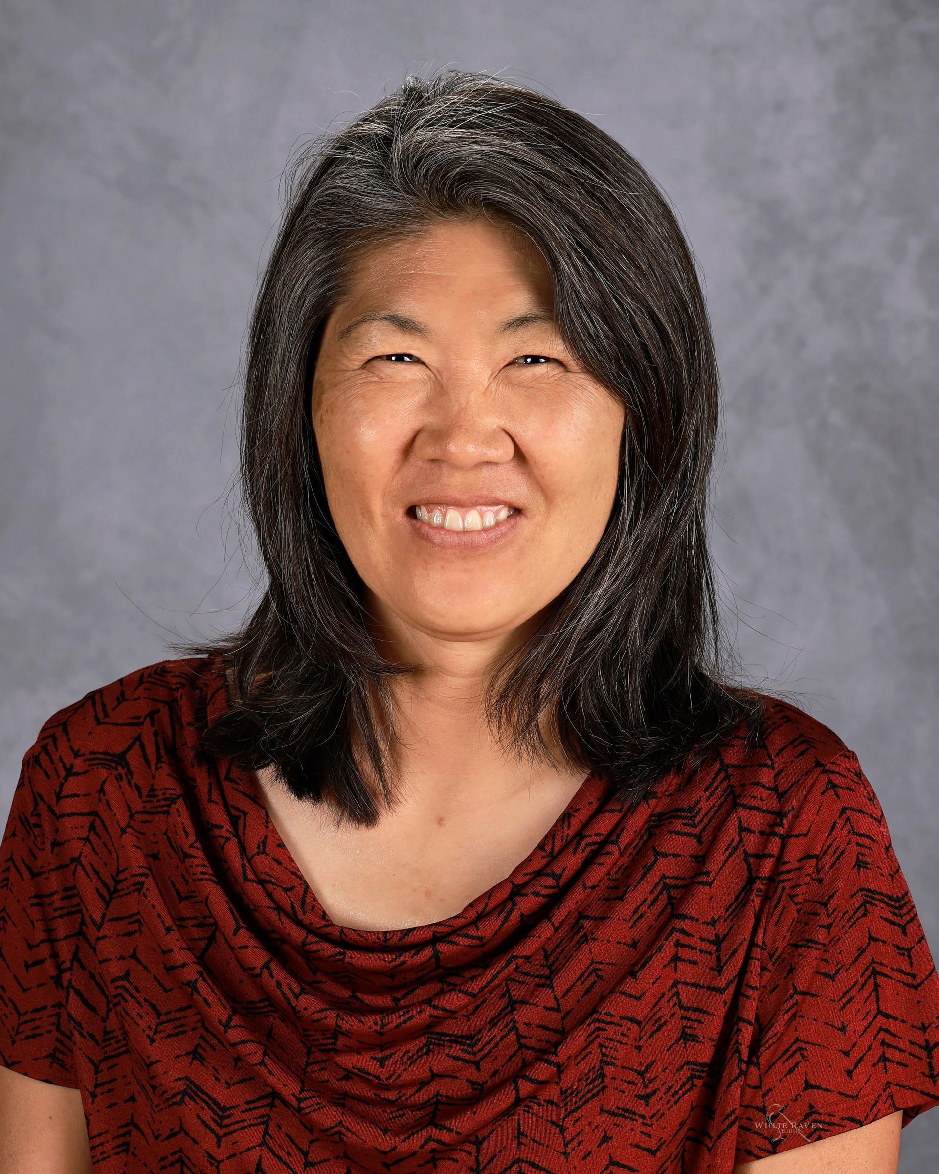 Woman with dark hair, wearing a red patterned top, smiling at the camera against a gray backdrop.