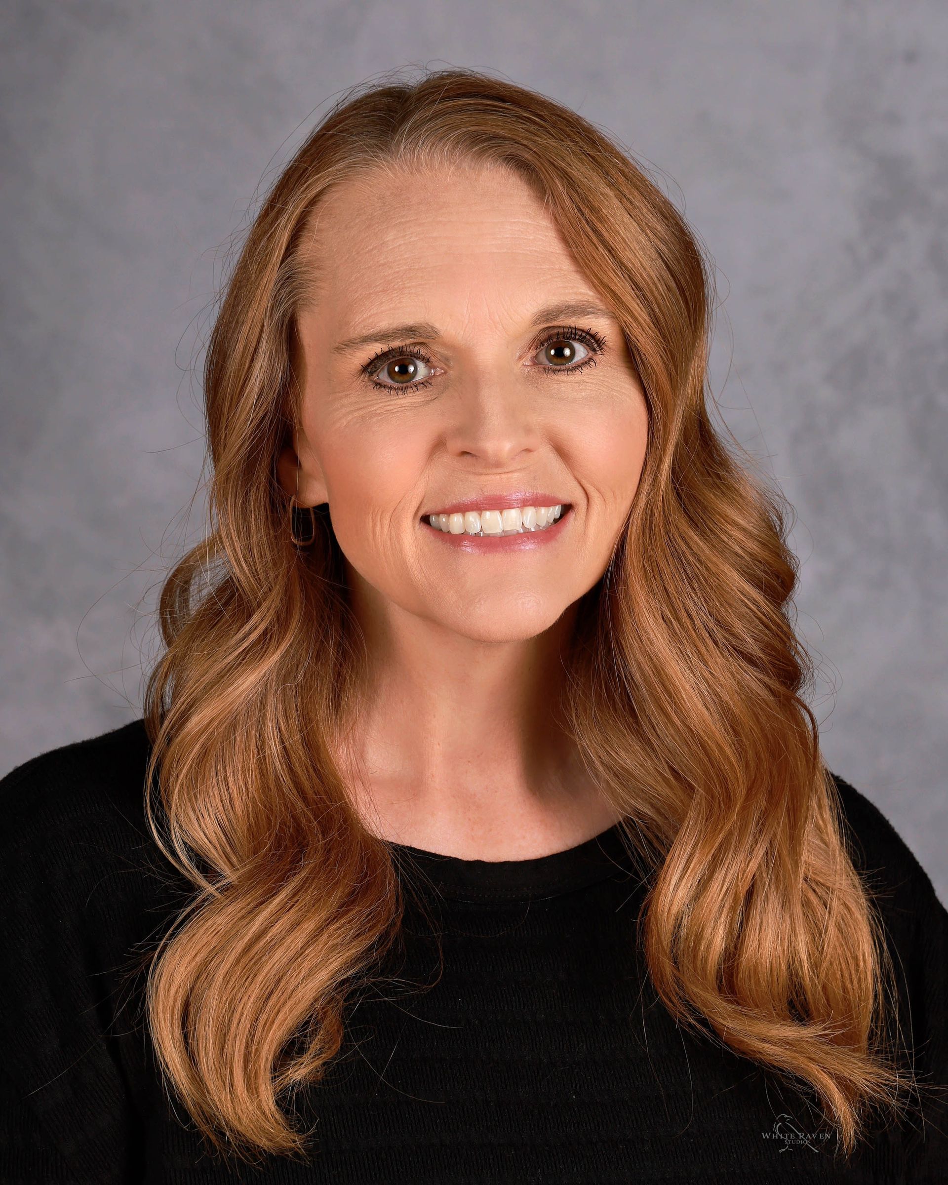 Woman with reddish-brown hair smiling; wearing a black top; against a gray backdrop.