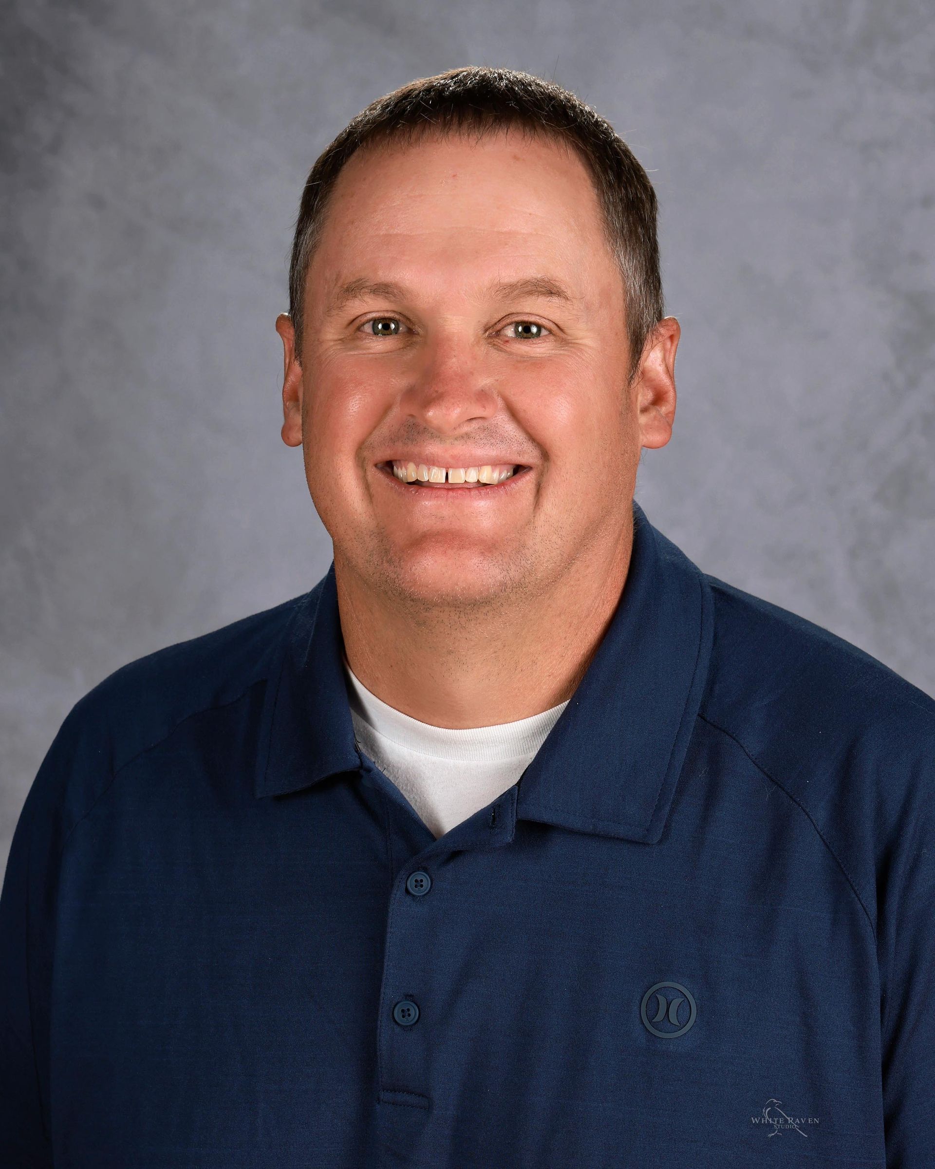 Smiling man wearing a blue polo shirt, white undershirt, and dark hair against a blurred gray backdrop.