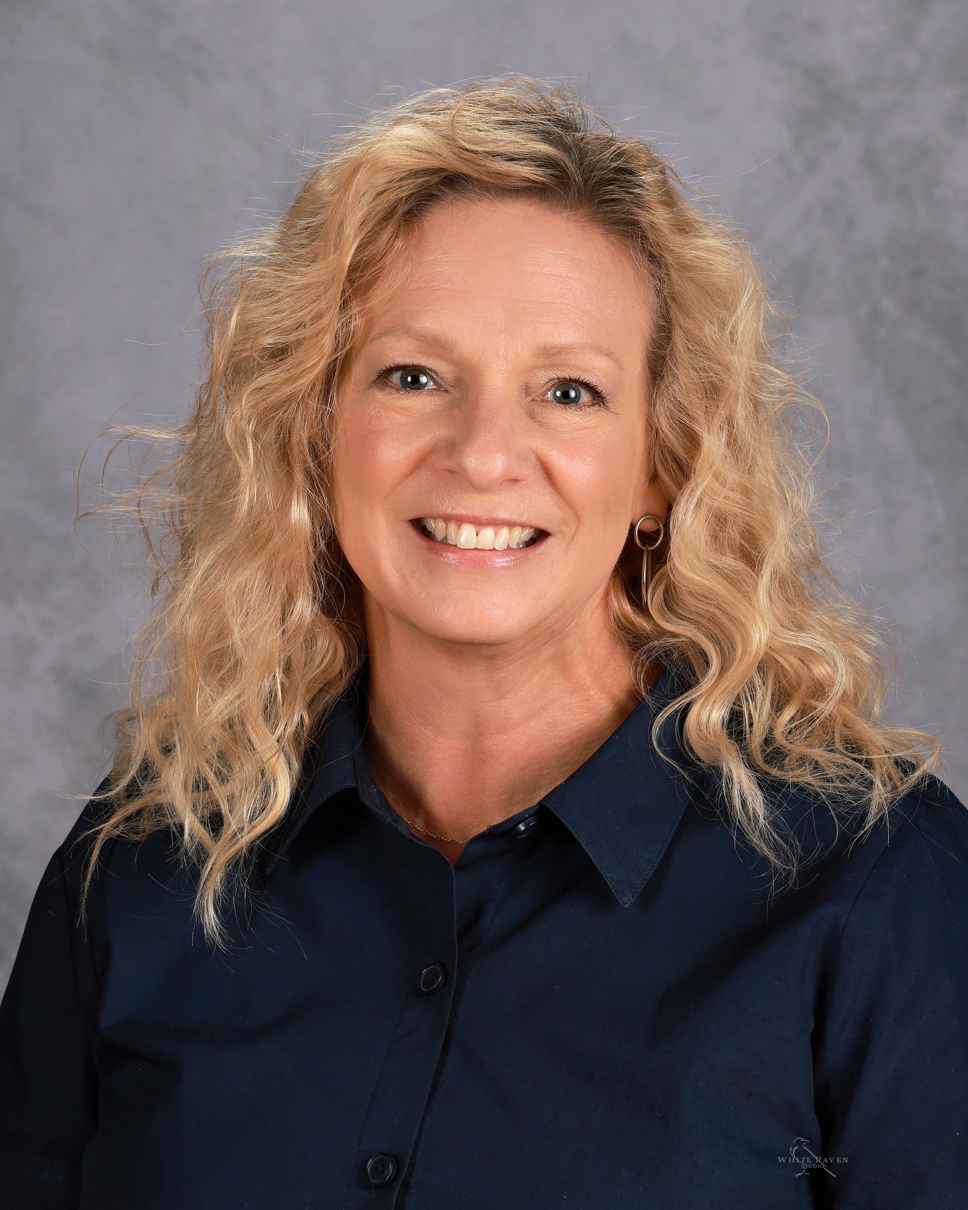 Woman with blonde curly hair smiles, wearing a navy blue button-down shirt, on a gray backdrop.