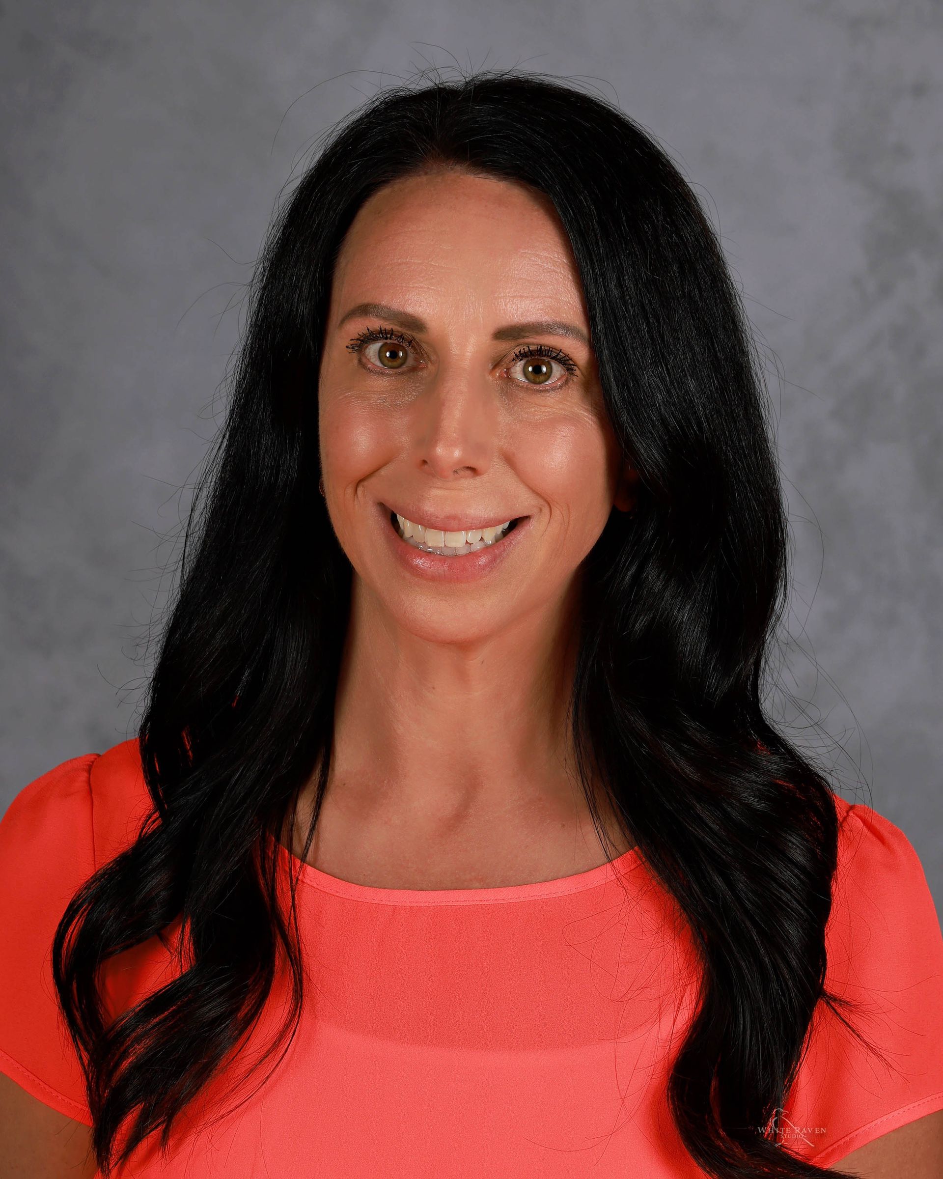 Woman with long black hair smiling in a coral shirt, against a gray background.