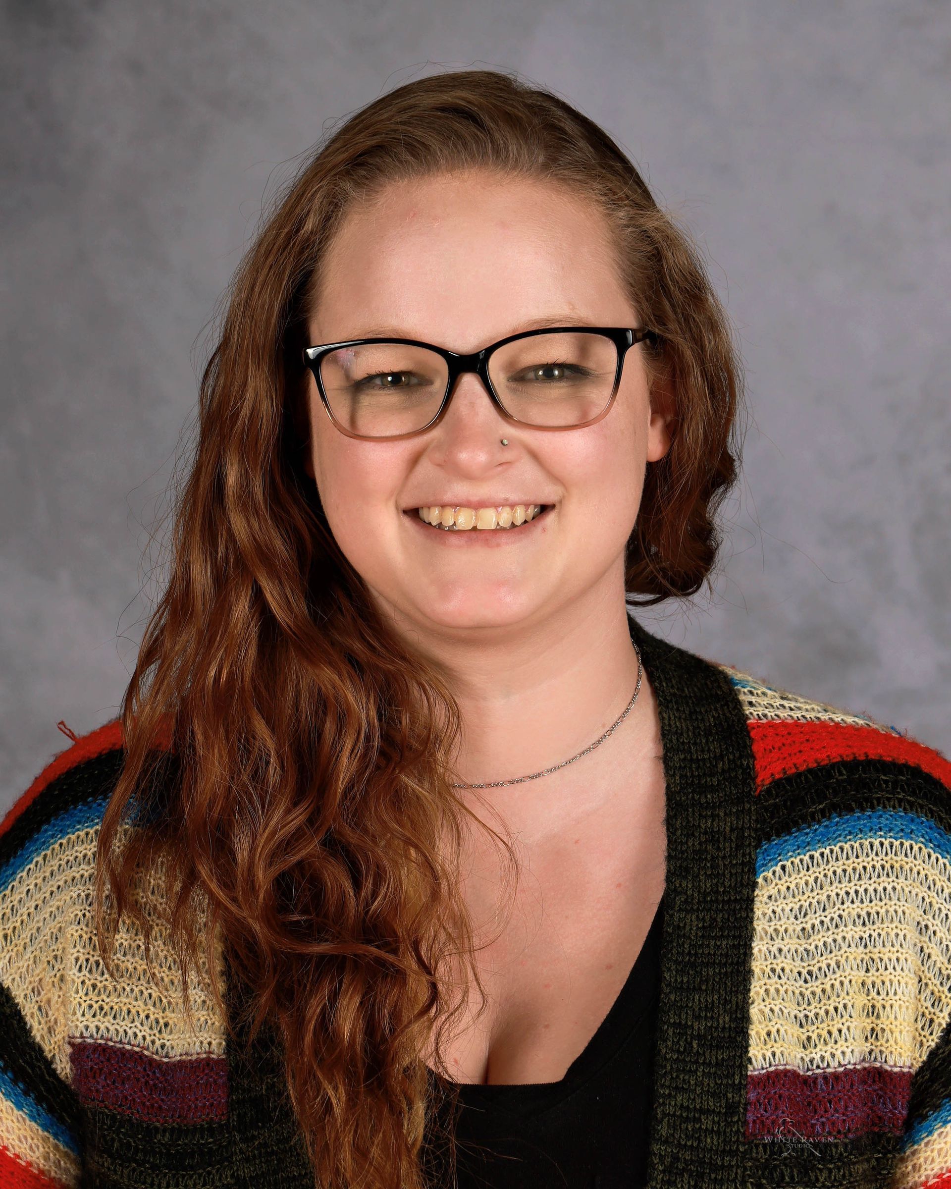 Woman with glasses and long red hair smiles, wearing a black top and colorful cardigan.