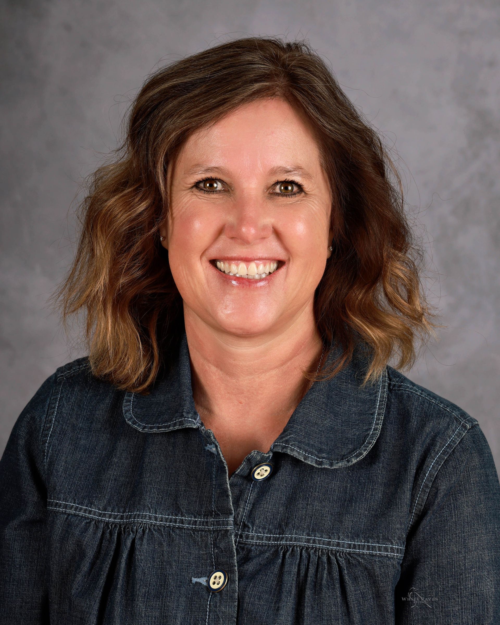 Woman with brown hair smiling, wearing a blue jean jacket against a blurred gray background.