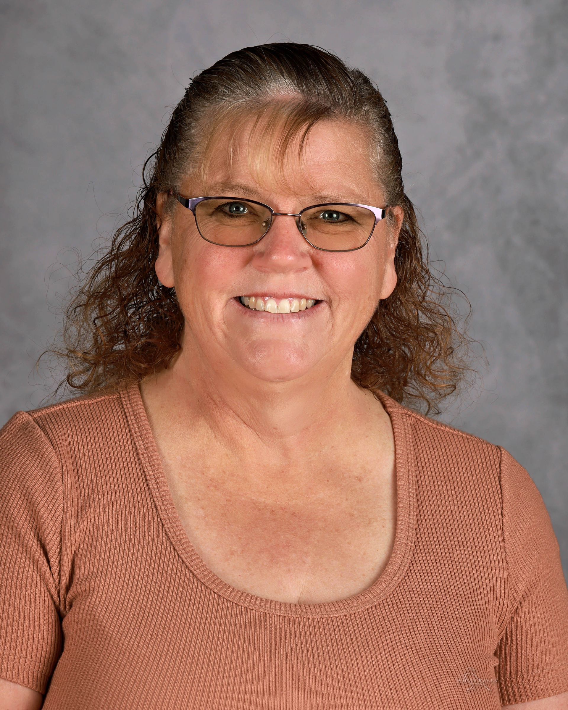 Woman with glasses smiling, wearing a pinkish-brown top, against a blurred gray background.