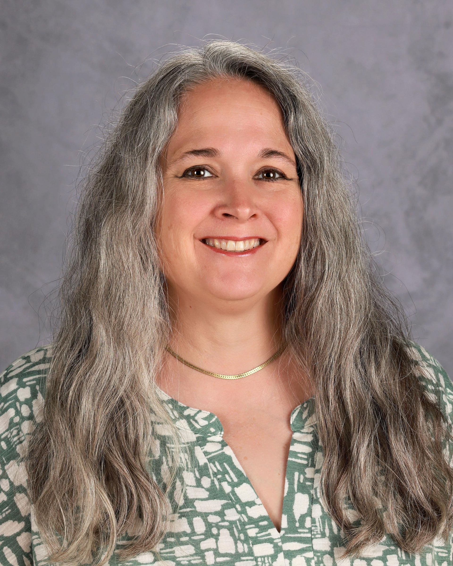 Woman with long gray hair, smiling, wearing a patterned green top and gold necklace.