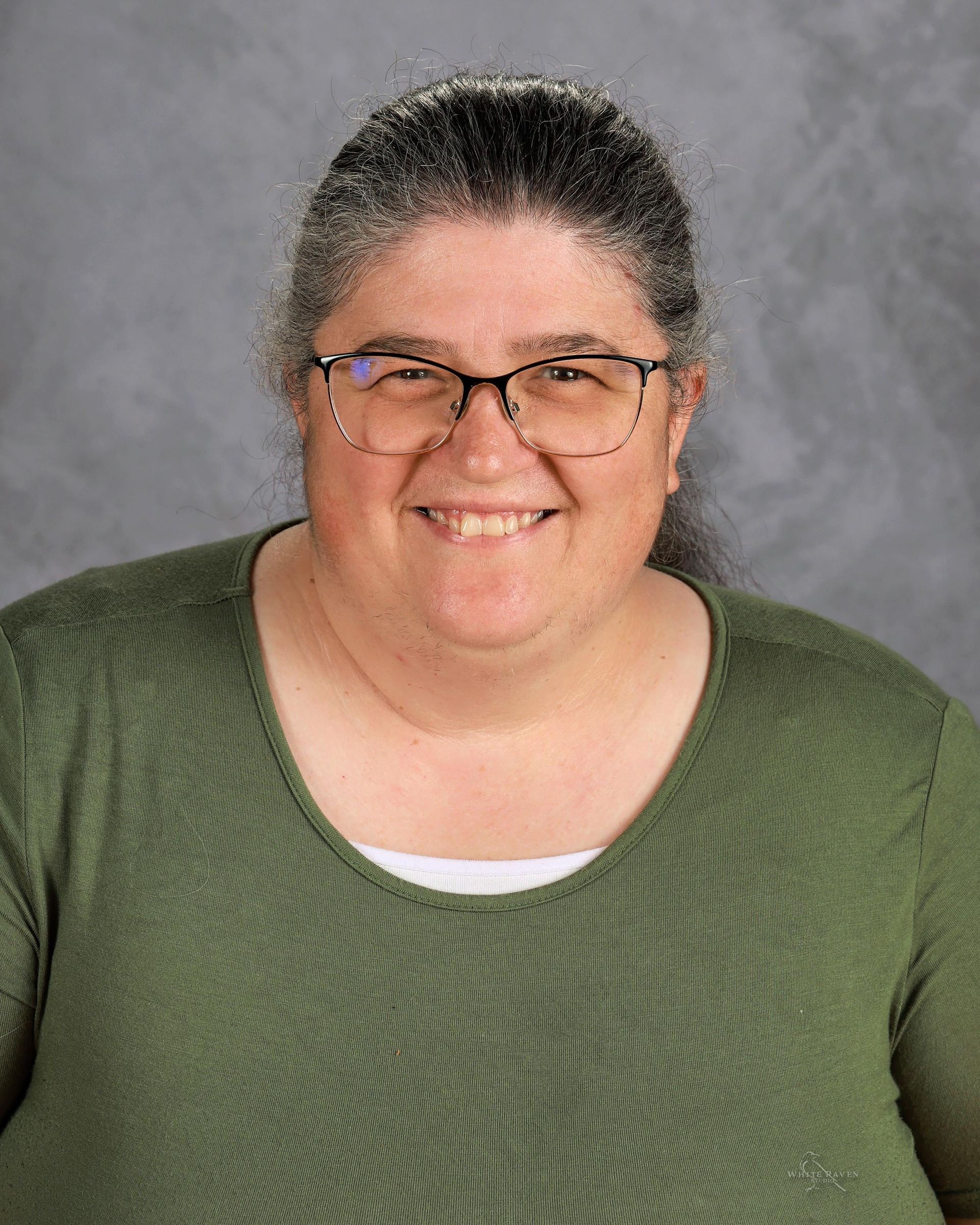 Woman with glasses smiles, wearing a green shirt, and posing in front of a plain background.