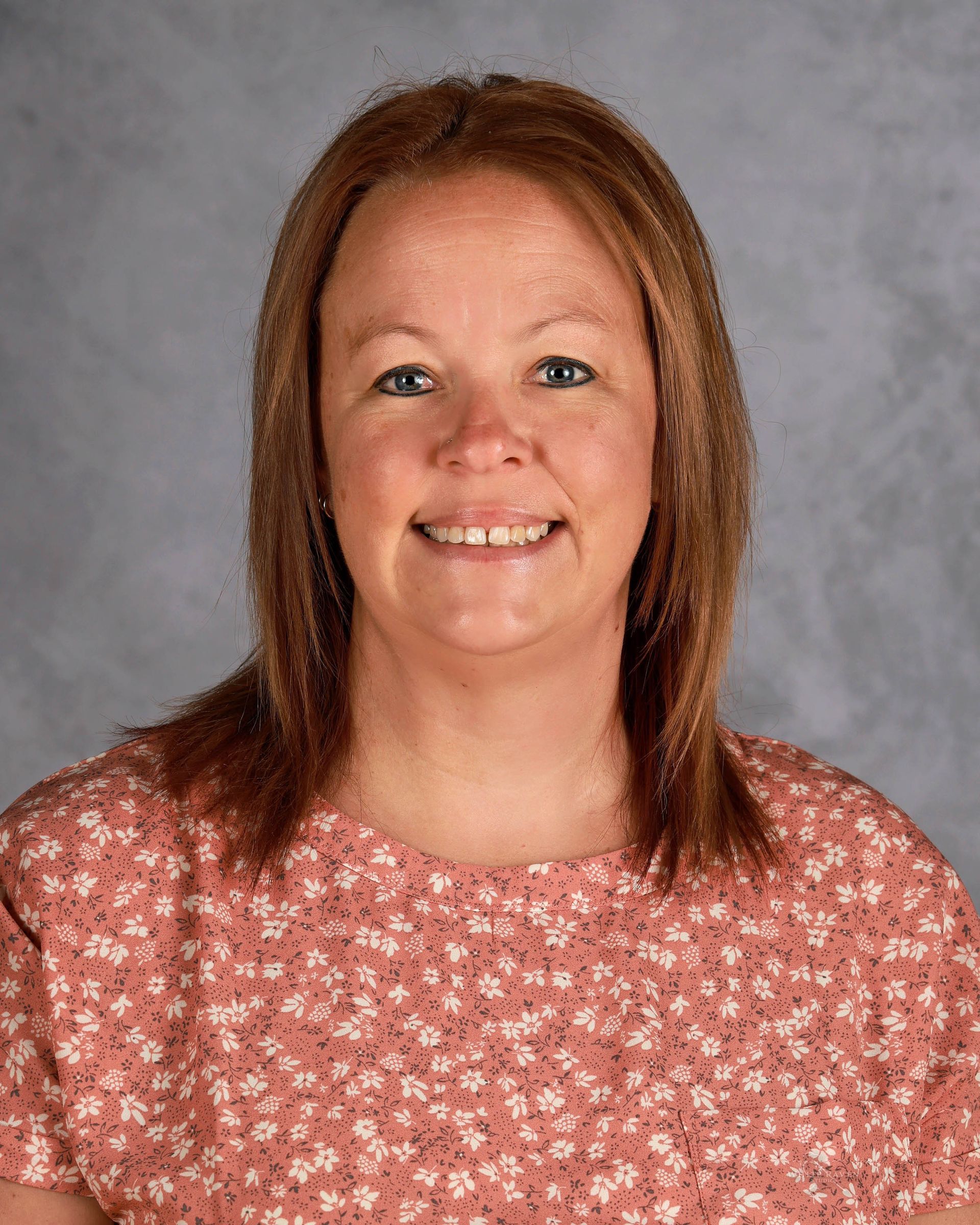 Woman with reddish-brown hair smiles, wearing a pink floral shirt, against a gray backdrop.
