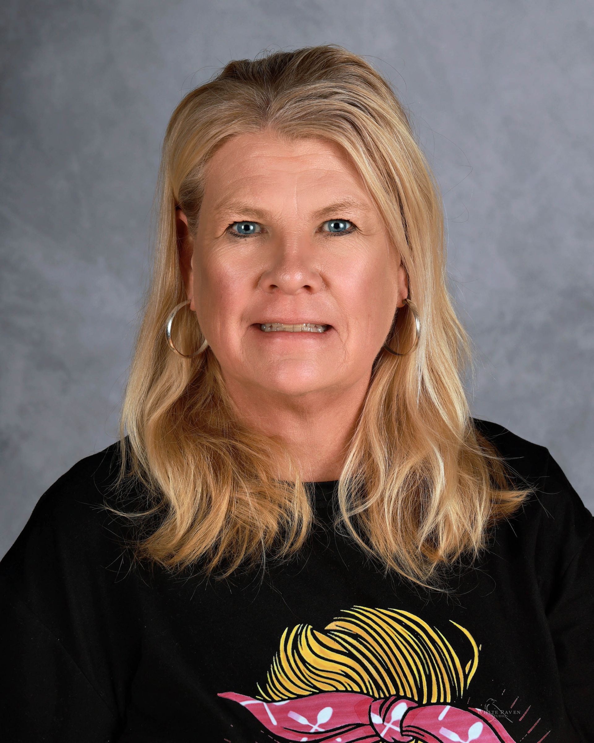 Woman with blonde hair smiles, wearing black shirt with a cartoon design, posed against a gray backdrop.