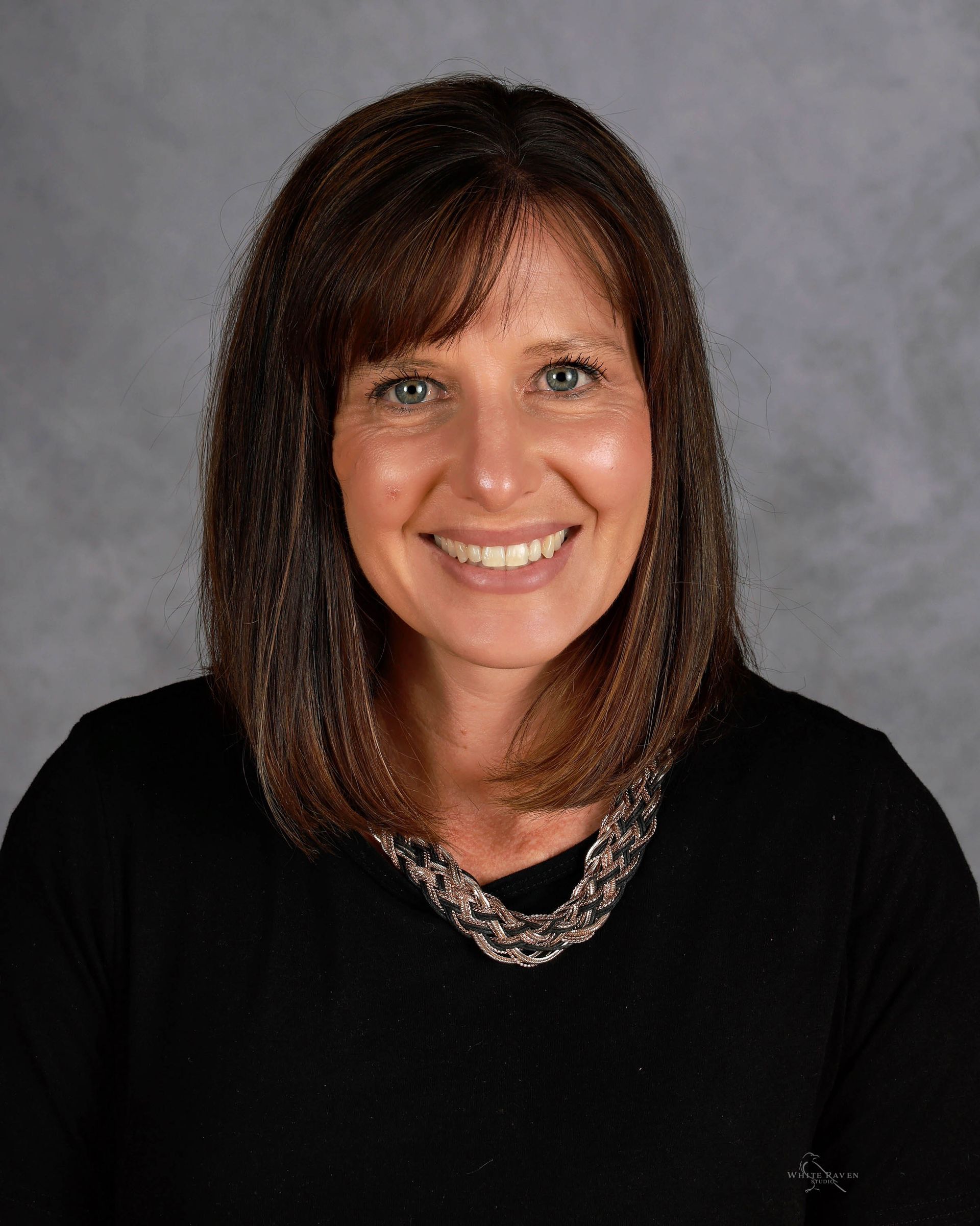 Woman with brown hair, smiling, wearing a black top with a decorative necklace. Gray background.